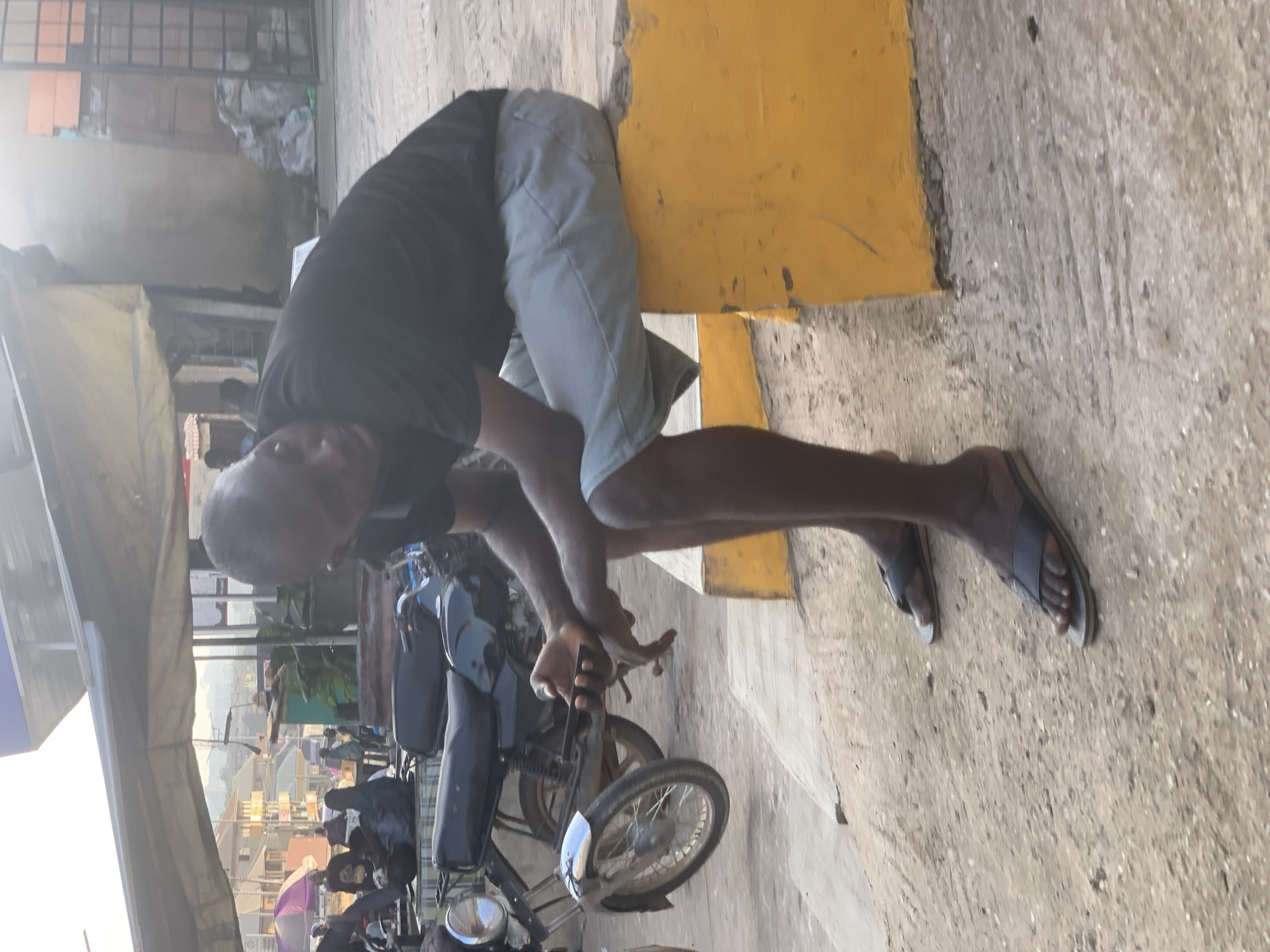 In Shagamu, Nigeria, a man is seated on a low, yellow, concrete barrier. He is wearing a black t-shirt and light-colored shorts, and dark-colored sandals. His head is bowed, and he is looking down at his phone, which he is holding in his right hand. To his left, a motorcycle is parked. In the background, there are buildings, a tent-like structure providing shade, and other people. The ground is made of dirt and concrete. The lighting suggests it is daytime, possibly late afternoon.