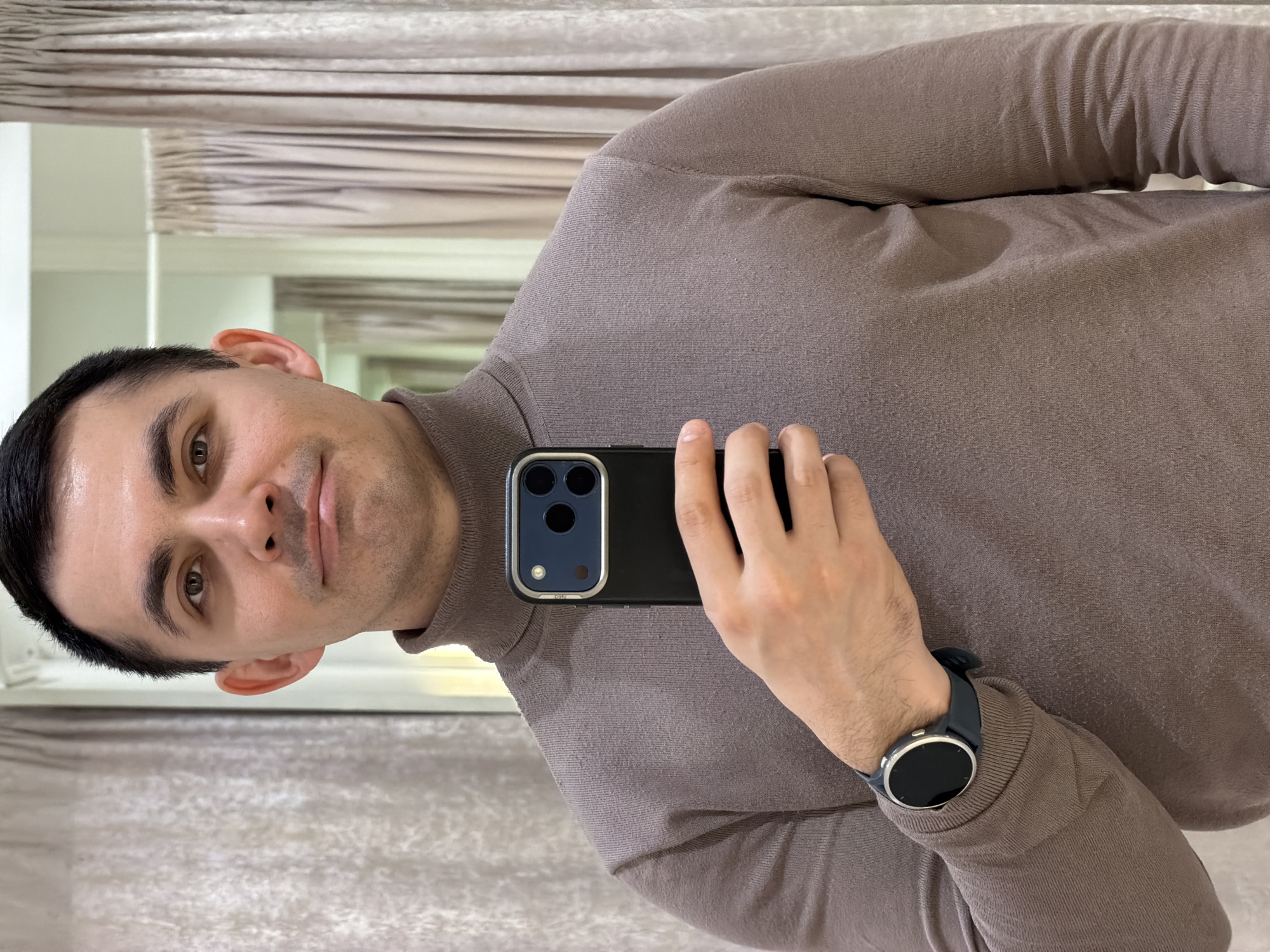 A man with dark hair is taking a selfie in a room with neutral-toned curtains or blinds. He is wearing a brown turtleneck sweater and a smartwatch on his left wrist. He is holding a black phone with multiple camera lenses. The room has mirrors that reflect parts of the room. There is no indication of the location in Izhevsk, Russia.