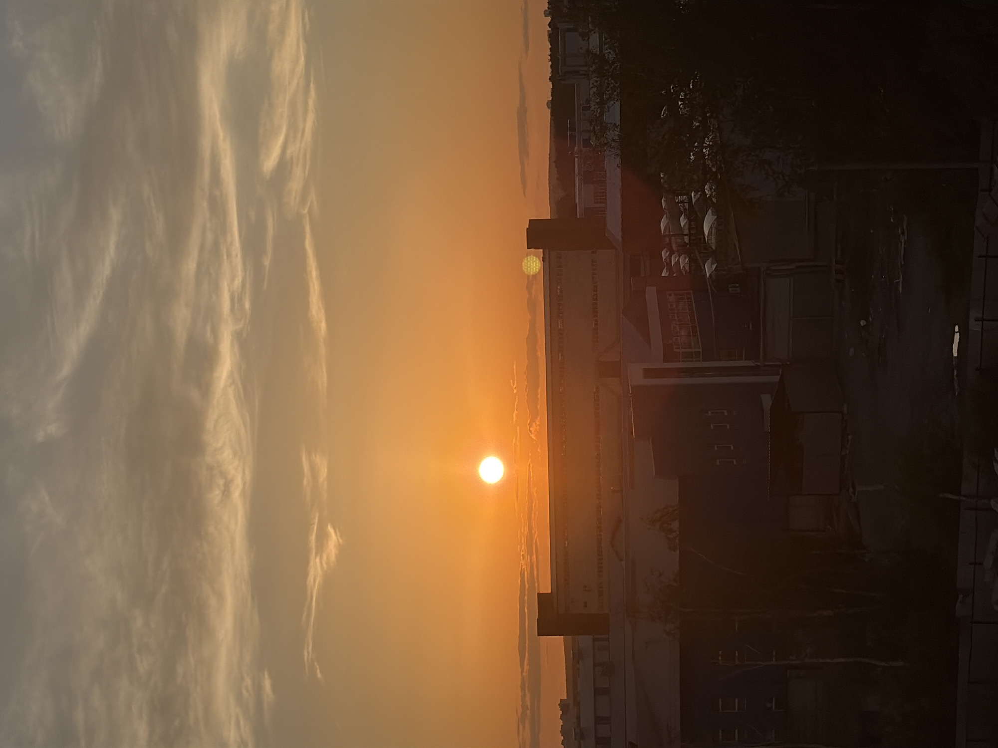 The image depicts a city landscape during a sunset in Yekaterinburg, Russia. The sun, a bright orb of orange, is low on the horizon, casting a warm glow across the sky and illuminating thin, wispy clouds. Below the sky, a series of buildings, likely industrial or commercial structures, are visible. These buildings vary in height and architectural style, with some featuring large facades and others appearing as lower structures. The foreground is darker, showing trees and hints of more urban infrastructure. The overall impression is one of an industrial area at the end of the day, bathed in the atmospheric light of sunset.