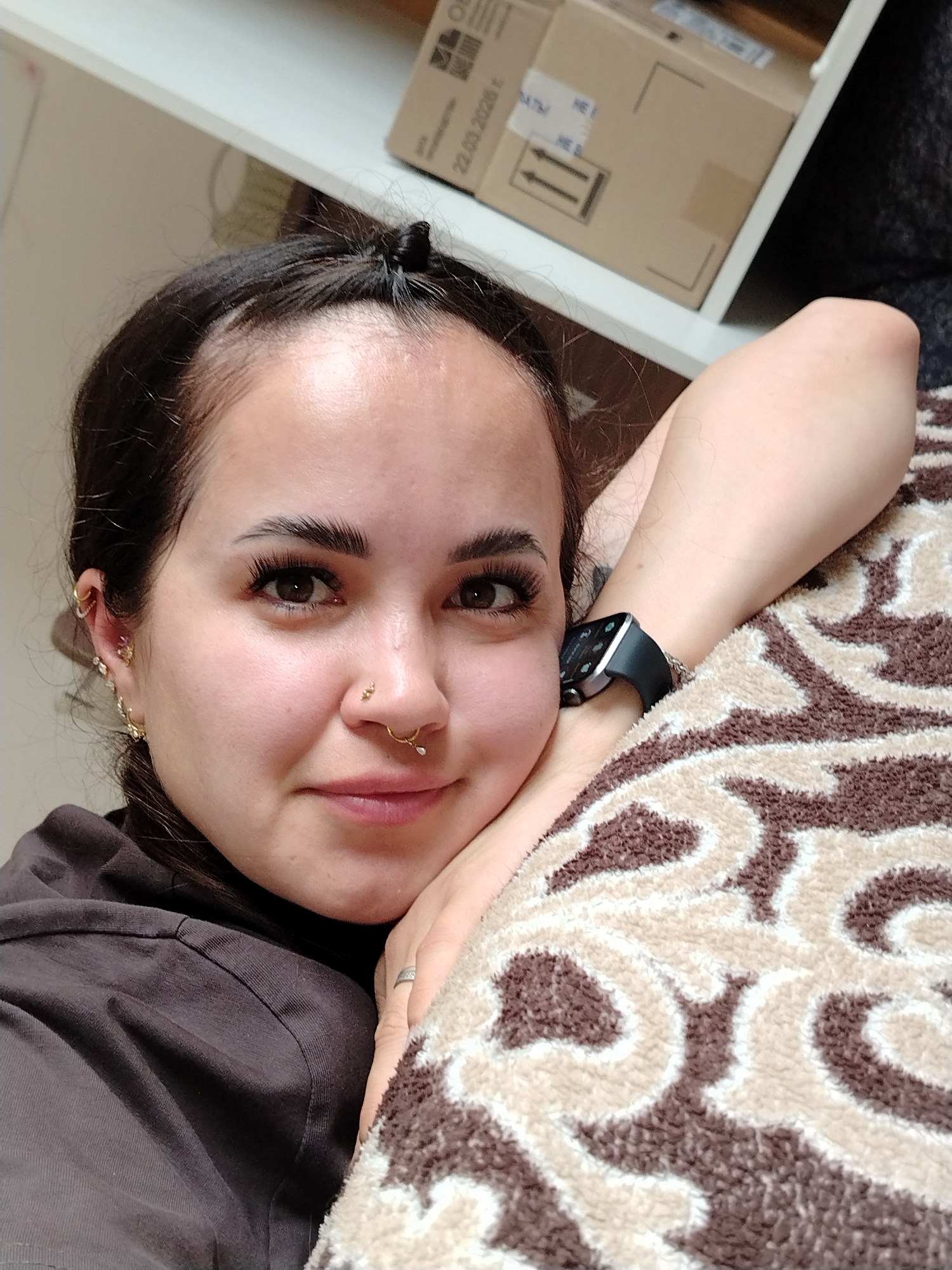 In an indoor scene, a young woman with dark hair pulled back in a bun rests her head on her arm, which is covered by a patterned blanket. She is wearing a dark shirt and has several piercings, including in her ear and nose. A smartwatch is visible on her wrist. In the background, a white shelf holds cardboard boxes. The scene takes place in Noviy, Russia.