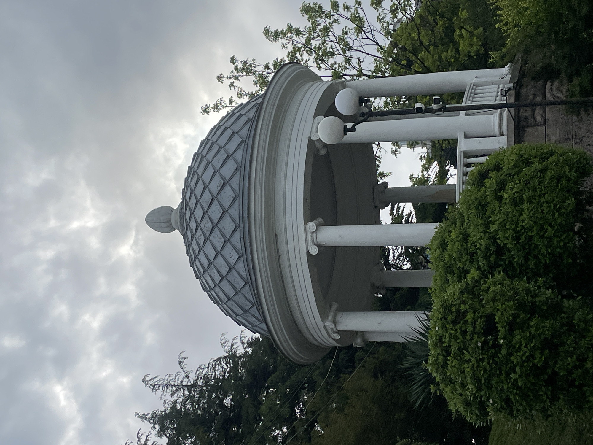 The image depicts a white, dome-topped gazebo with a tiled roof and white pillars, set against a cloudy sky and surrounded by greenery. Two spherical lampposts are attached to the gazebo's top structure. Surveillance cameras are mounted on the structure near the lampposts. The location is Sochi, Russia.