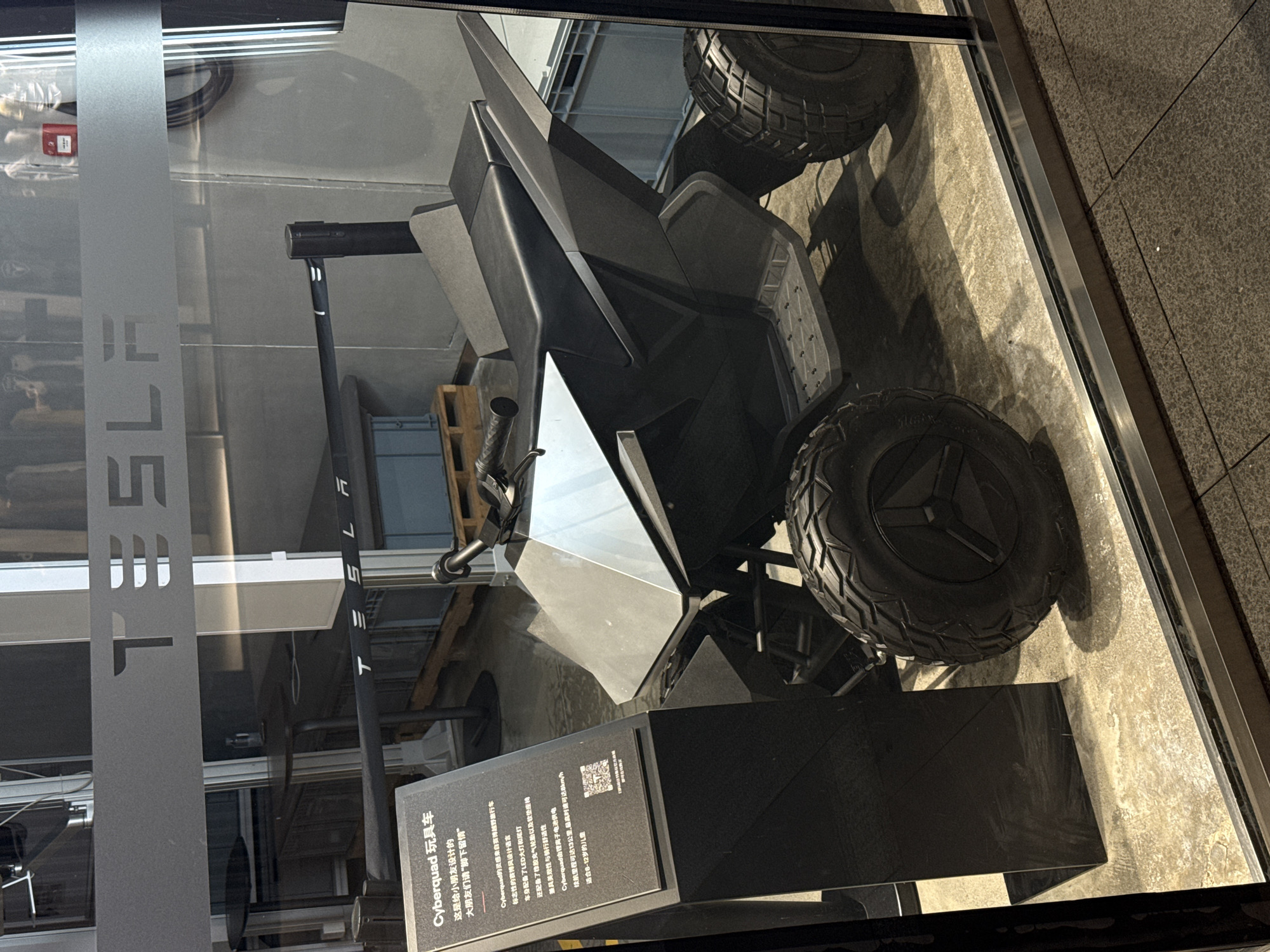 Here's a forensic description of the image:
The image shows a Tesla Cyberquad toy car displayed behind a glass window. The window is marked with the Tesla logo. The Cyberquad is dark gray with a futuristic design and large, knobby tires. A black sign in front of the vehicle provides information in Chinese, identifying it as a "Cyberquad toy car" designed for children aged 8-12. The sign states that it is inspired by the Cyber Truck and has LED headlights. The location is Jinjiang District, China. The image appears to be taken from outside the window, looking into a retail space. The exterior appears to be a concrete walkway.