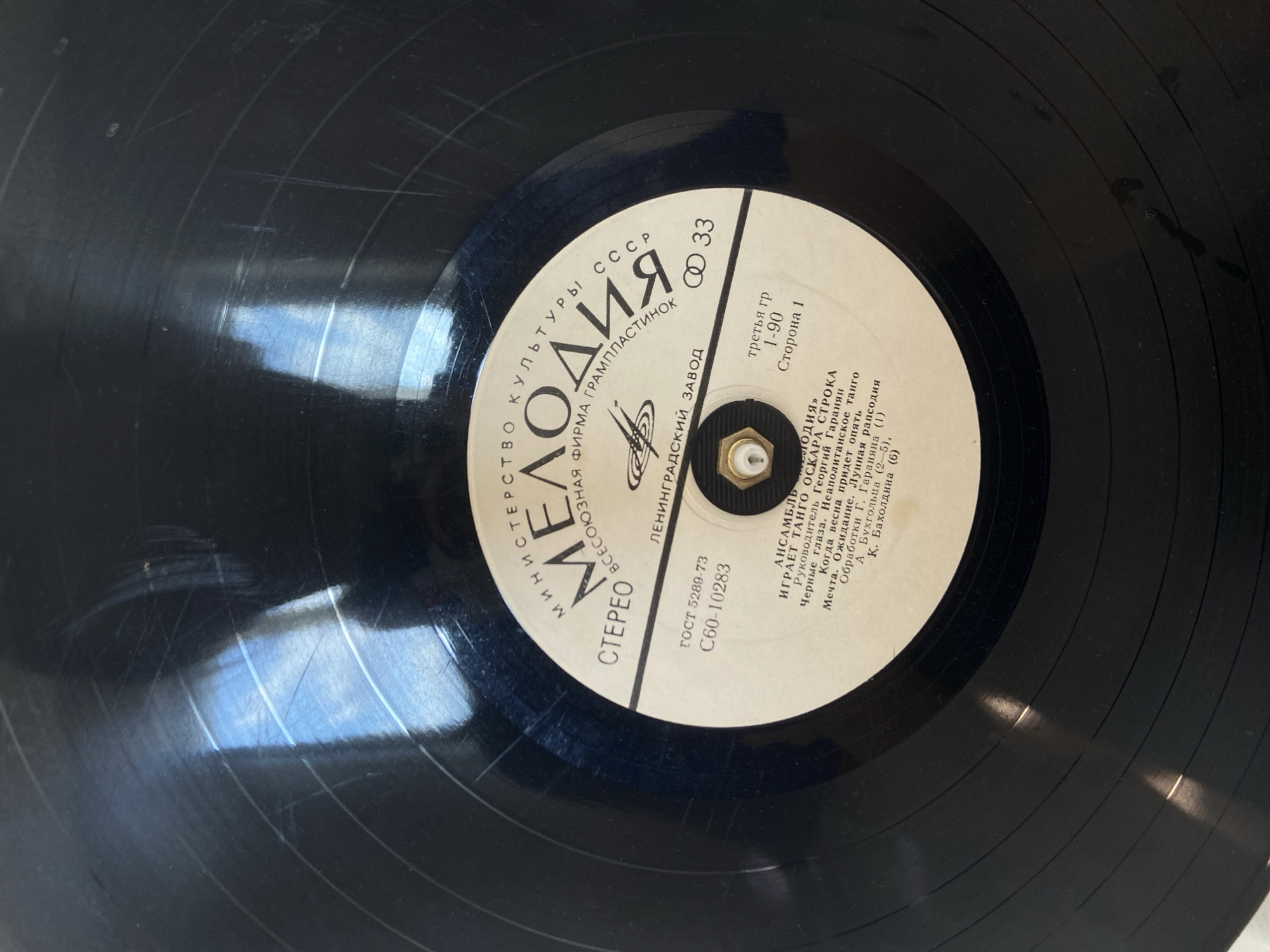The image is a close-up, eye-level, medium shot of the label of a black vinyl record, with its center hole clearly visible. The label is primarily white with black text and some circular graphics. The text on the label is in Russian and includes "МИНИСТЕРСТВО КУЛЬТУРЫ СССР," "МЕЛОДИЯ," "ВСЕСОЮЗНАЯ ФИРМА ГРАМПЛАСТИНОК," "ЛЕНИНГРАДСКИЙ ЗАВОД," and other song titles and credits. There are also markings indicating "ГОСТ 5289-73," "C60-10283," and "33" with a symbol. The record itself shows concentric grooves typical of vinyl records, and there are reflections of light on its surface, indicating a polished material. The overall environment suggests an indoor setting where the record is being presented or stored. There is no indication of the city of Krasnodar, Russia, or any people, actions, or interactions within the image.