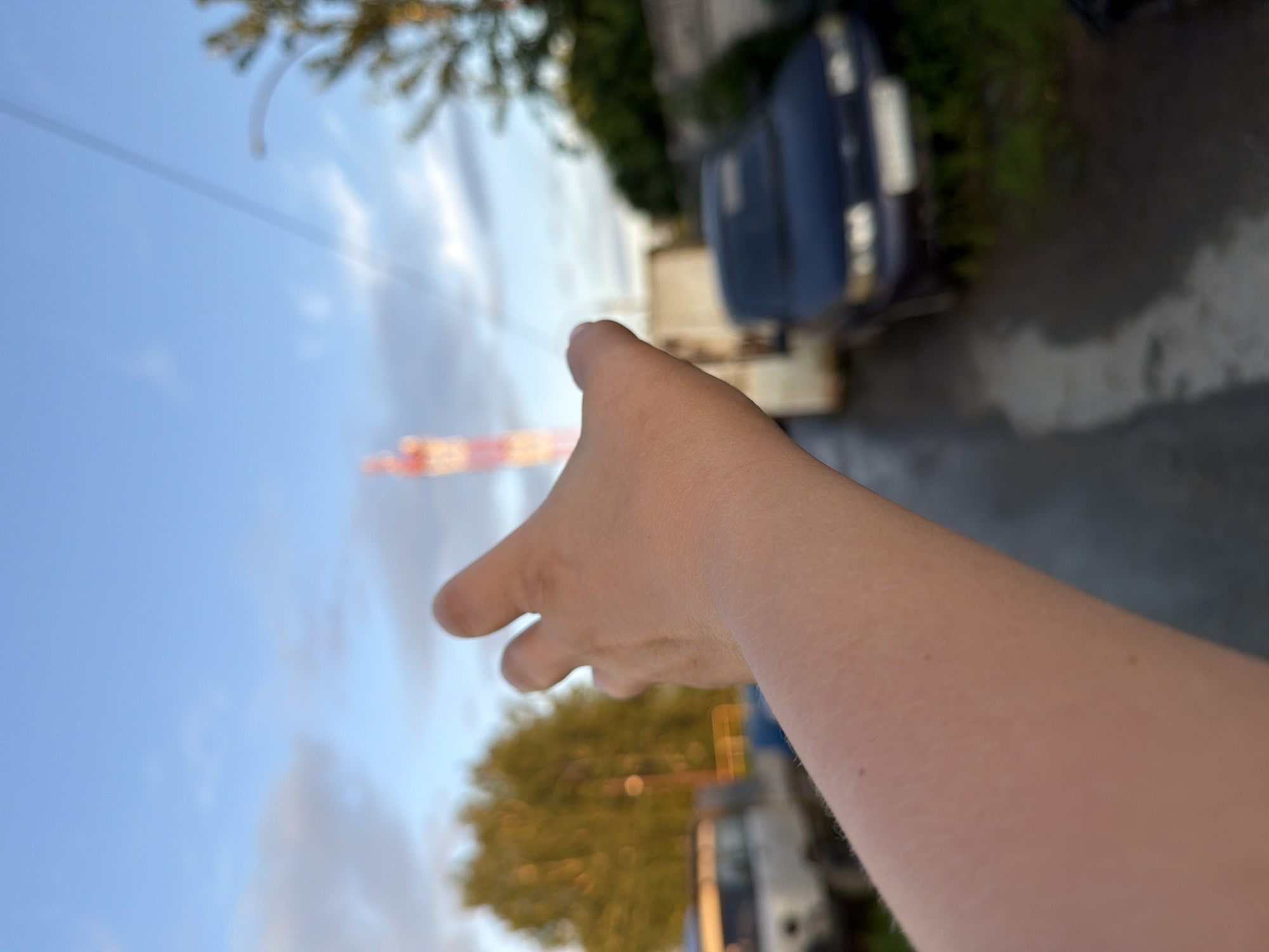 The image shows a person's arm and hand reaching out towards the sky. The hand is in the foreground, blurred and positioned to the right of the frame, with the fingers slightly curled as if grasping for something. The arm extends out of the right side of the image.
In the background, the scene is a sky with some clouds and the tops of trees. There is a thin, dark line that could be a power line or a wire running horizontally across the sky. In the distance, a blurry object appears to be a tower or a structure with horizontal bands of orange and yellow, suggesting it might be a construction crane or a radio tower.
To the left of the hand, a dark blue car is visible, parked on an asphalt surface. Green foliage is present behind the car, indicating an outdoor setting. The lighting suggests it is daytime, possibly late afternoon or early evening due to the soft light. The overall mood is somewhat aspirational or longing, with the extended hand reaching towards the distant structure and the open sky. There is no visible text in the image.