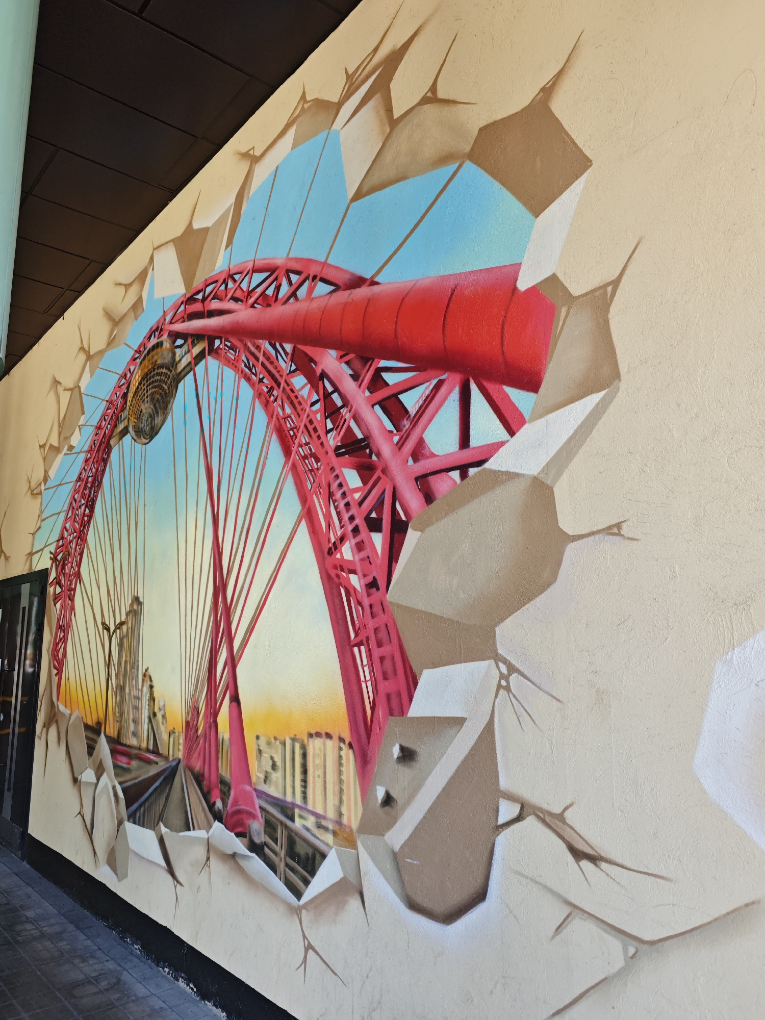The image depicts a vibrant mural painted on a wall, creating a 3D illusion of a bridge bursting through. The mural showcases a prominent red cable-stayed bridge, likely the Zhivopisny Bridge in Moscow, Russia, with its distinctive arch and radiating cables. The background features a bright blue sky with hints of an orange sunset, and a cityscape of buildings in the distance. The mural is designed to appear as if pieces of the wall are breaking away, revealing the bridge and sky behind. The perspective suggests the viewer is looking at the mural from a slightly low angle. The foreground shows the textured surface of the wall with painted cracks and shadows, enhancing the 3D effect. The bottom of the image shows a tiled walkway and a black baseboard.