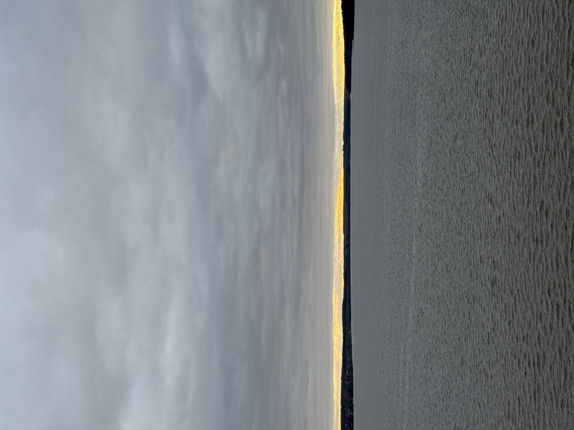 The image presents a landscape with a body of water dominating the right side. Its surface appears rippled. On the left side, the sky is covered by dense, gray clouds. A strip of land, barely visible, separates the water and the sky. A thin, bright band, likely sunlight filtering through the clouds or reflecting off the water, illuminates the edge of this land. The image does not contain discernible people, subjects, or interactions. The environment suggests an outdoor setting, but specific location is not identifiable from the image.