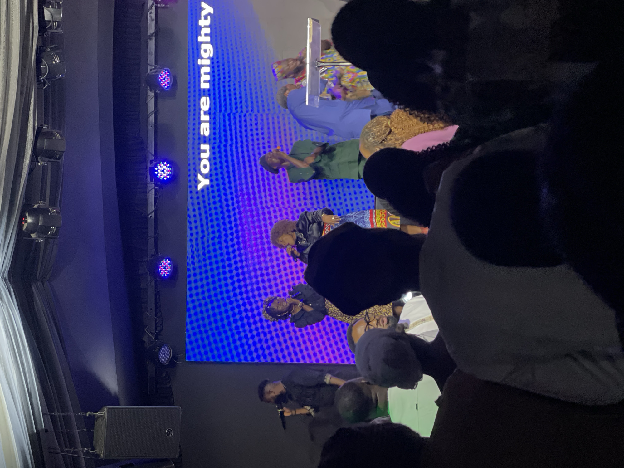 A dimly lit indoor stage in Jos, Nigeria, featuring a digital screen displaying the words "You are mighty" in white text against a blue patterned background. On the stage, a group of performers sing into microphones. The foreground is filled with an out-of-focus audience, partially obscuring the view. Stage lighting fixtures and speakers are visible on the left side of the frame.