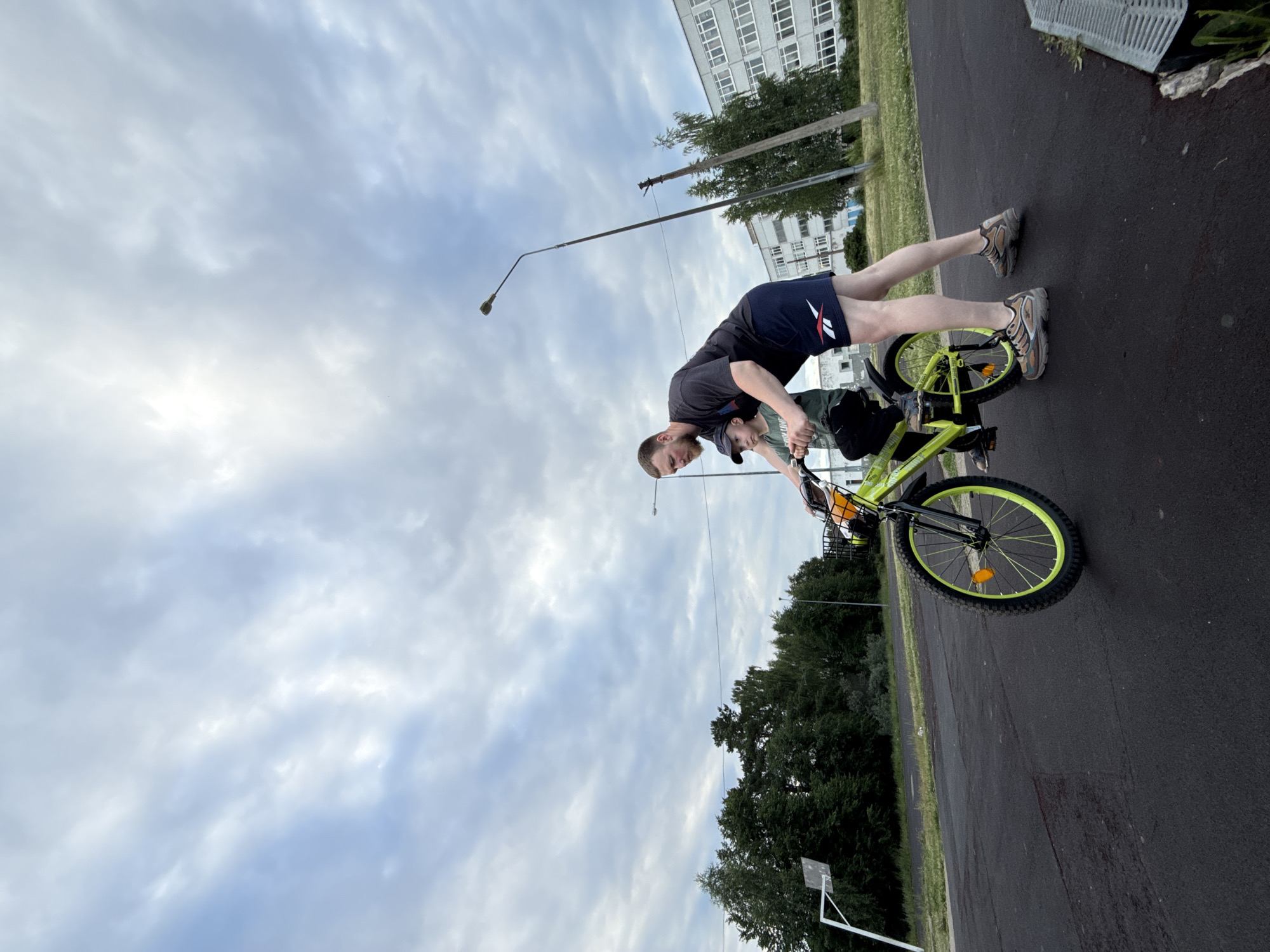 In the city of Chaikovsky, Russia, on a cloudy day, a man is assisting a young boy in riding a bright green bicycle on an asphalt-paved area, possibly a sports ground. The man, wearing a dark t-shirt and blue athletic shorts, is leaning over and holding onto the bicycle's handlebars. The boy, dressed in a green shirt and a baseball cap, is seated on the bicycle and appears to be pedaling. The bicycle has a basket at the front with an orange reflector. In the background, there is a dense line of green trees, a basketball hoop structure, a tall lamppost, a utility pole, and a light-colored, multi-story building. The sky is overcast with dramatic clouds.