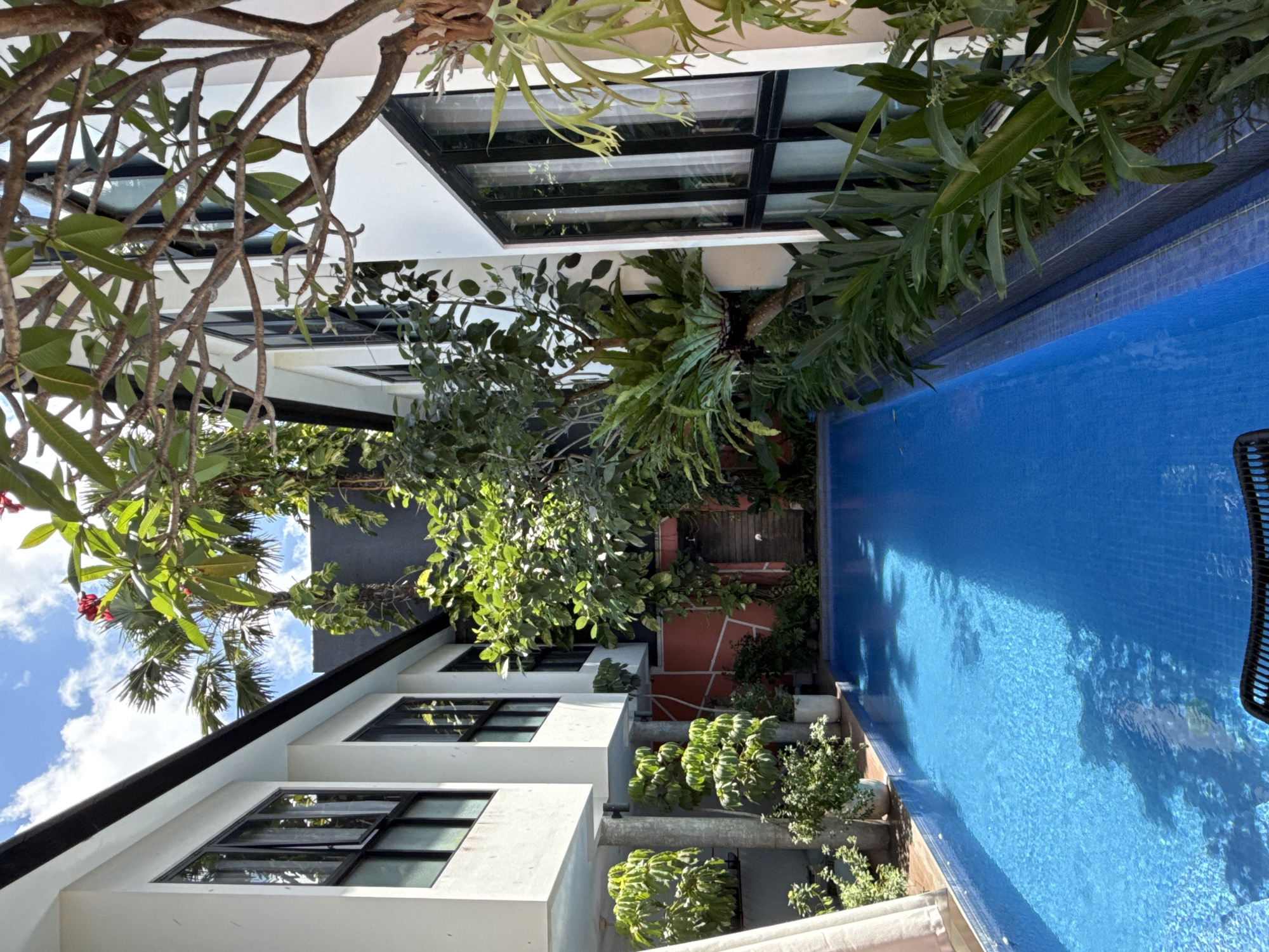 The image depicts a residential area in North Kuta, Indonesia. It shows a swimming pool next to white, modern-style buildings with black-framed windows. There is ample vegetation including trees and other plants, partially obscuring the view of the buildings and pool. The sky is visible in the background with some clouds.