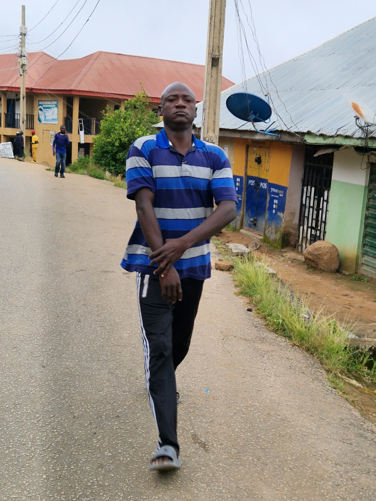 A bald man in a blue and grey striped polo shirt and black pants with white stripes walks toward the camera on an asphalt road. He has his arms crossed in front of him. In the background, there is a light-colored building with a reddish-brown roof, a yellow building with a blue satellite dish on its roof, and a green building with a metal grate. There are several power lines and a utility pole visible. Green grass and plants grow along the side of the road.