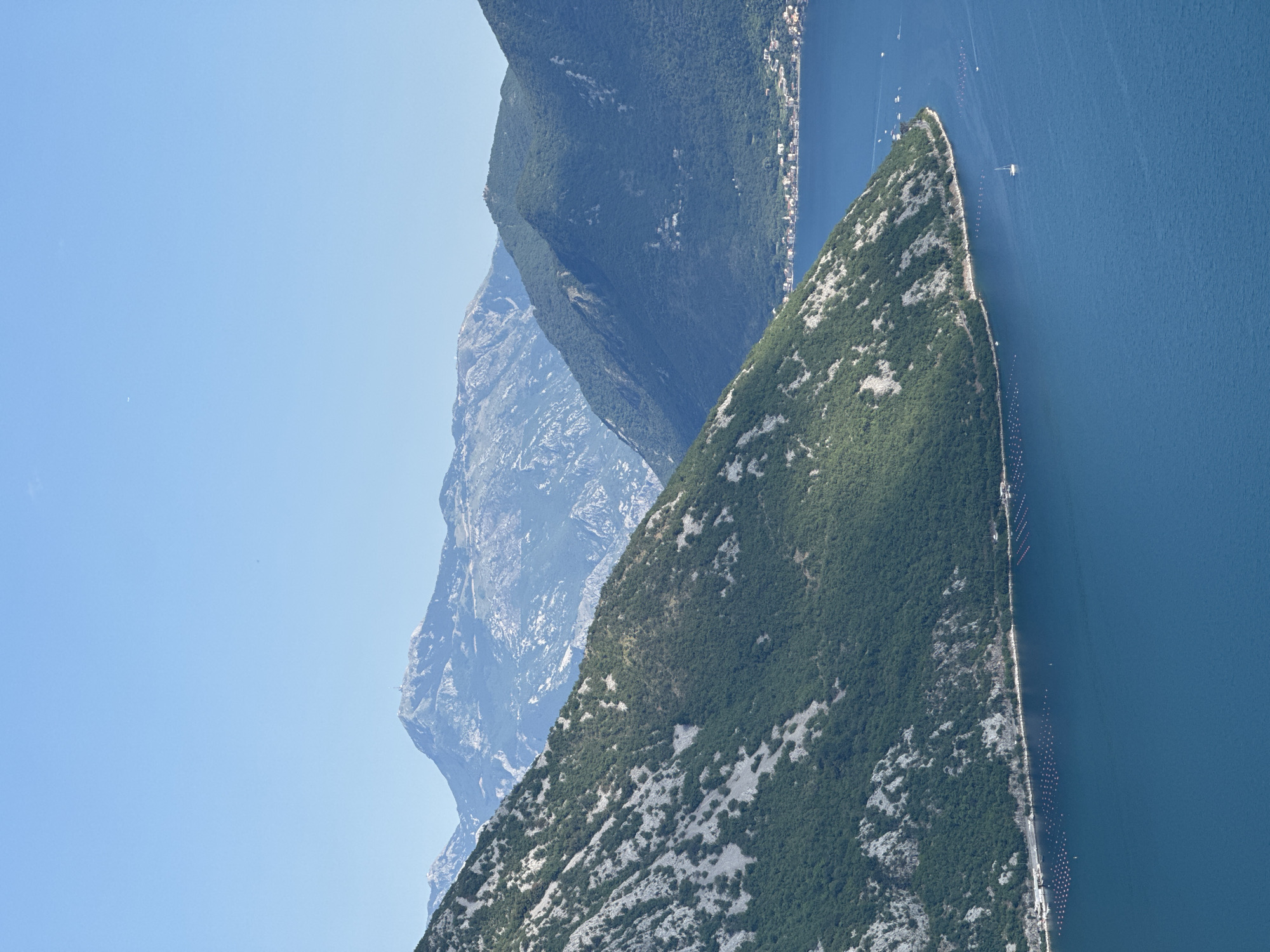 This is a vertical, eye-level, medium shot taken from a high vantage point overlooking a body of water. The water, a deep blue, takes up the bottom half of the frame. In the foreground, a steep, rocky mountain slope covered with green vegetation dominates the left side of the image. The slope curves towards the water, where it meets a shoreline. Along the shoreline, there are rows of buoys marking an aquaculture farm, and a few buildings of a town are visible in the distance on the right. Several small boats, including a sailboat, are scattered across the water. Further back, a series of progressively more distant mountain ranges stretch across the background, fading into a hazy blue under a clear, pale blue sky. The closest mountains are covered in dense green vegetation, while the furthest ones are rocky and appear snow-capped in some areas, possibly a distant view of Mount Lovcen. The scene appears to be of the Bay of Kotor near the town of Štrp, Montenegro.