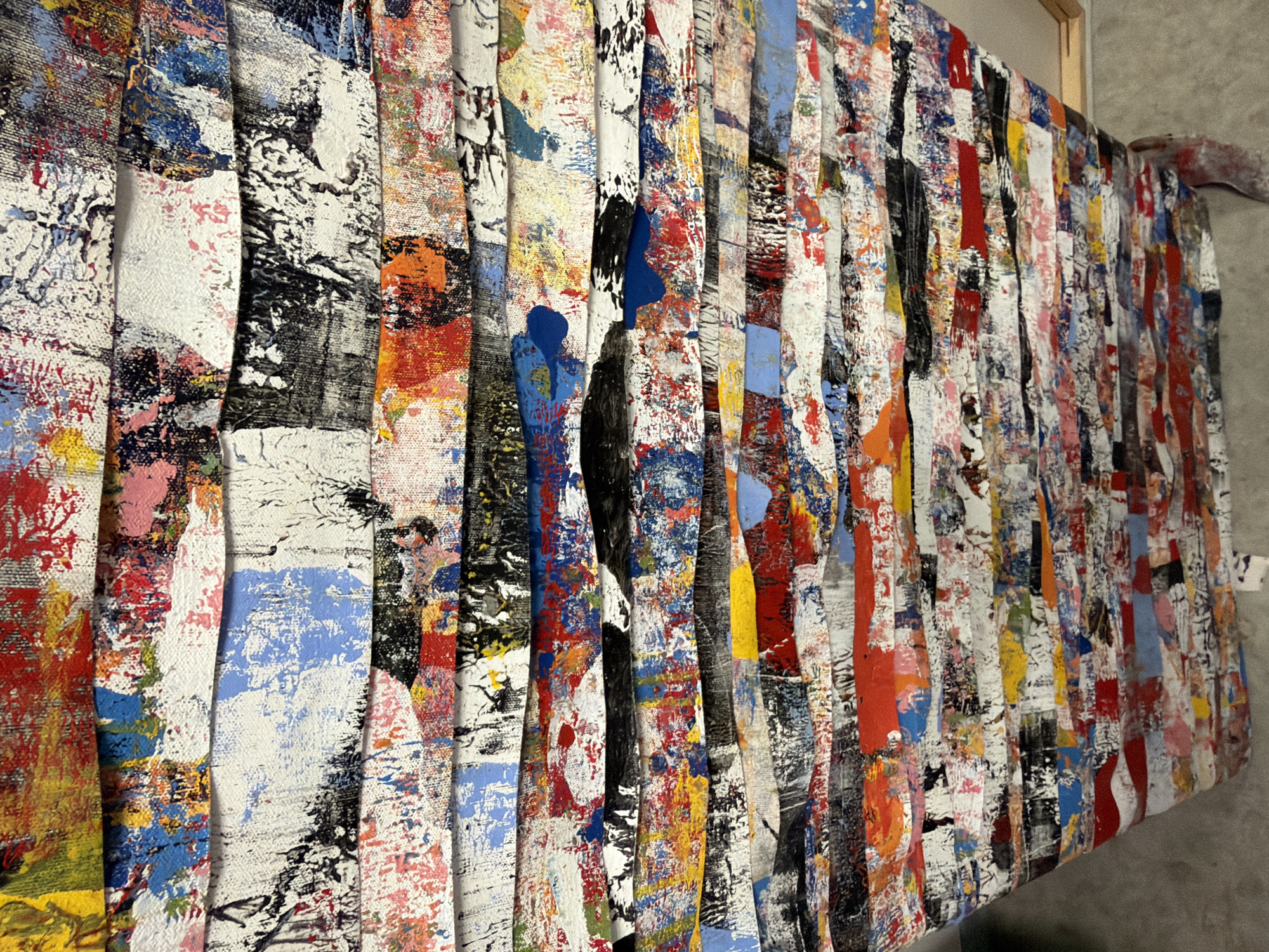 A close-up shot presents an abstract artwork composed of vertically arranged, narrow strips of canvas. Each strip is adorned with a vibrant and chaotic mix of paint splatters and brushstrokes in various colors including red, blue, yellow, orange, white, and black. The canvas strips are mounted onto a frame, visible at the top edge. The backdrop is a neutral gray wall. The image was most likely not taken in Toulon, France.