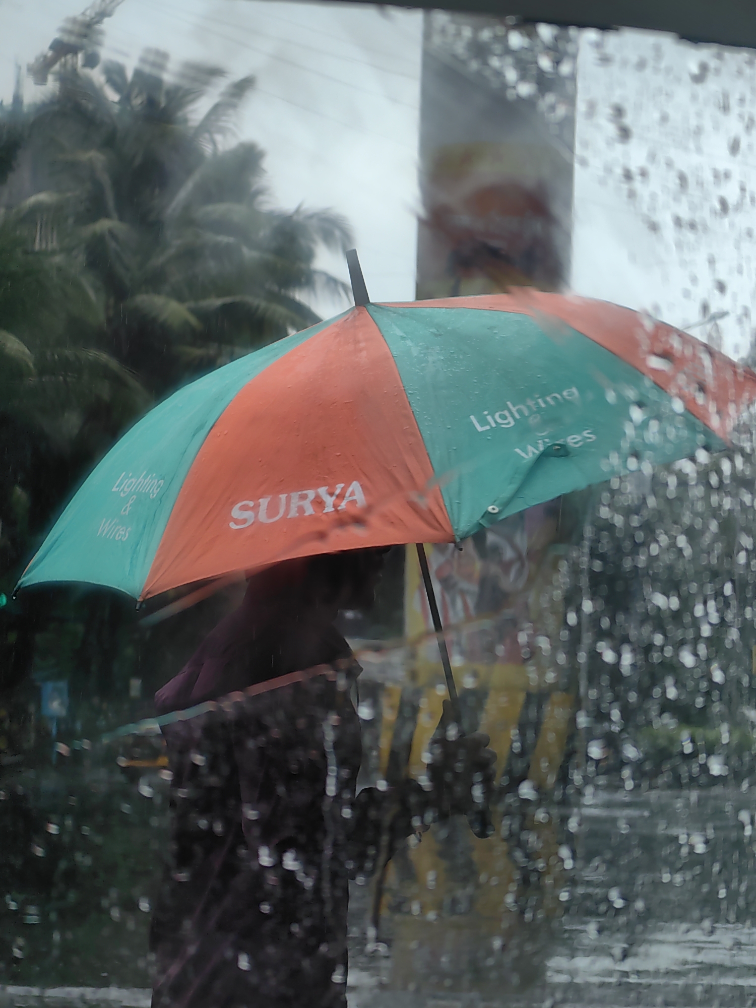 The image depicts a person holding an open umbrella with alternating orange and teal panels. The umbrella has "Lighting & Wires" written in white text on the teal panel, and "SURYA" written in white text on the orange panel. The scene is set during a rain shower, evidenced by raindrops on the window through which the photo is taken and the wet appearance of the umbrella. In the background, lush green palm trees are visible, suggesting a tropical or subtropical environment. The overall atmosphere is subdued due to the overcast weather. The location is identified as being in Mumbai, India.