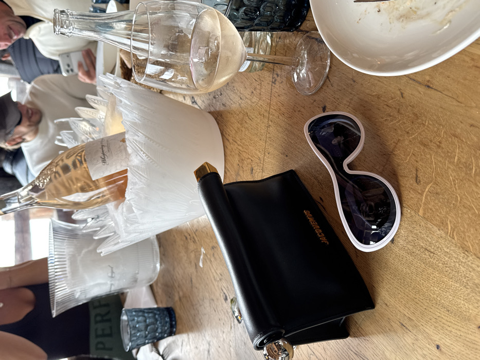 The scene captures a dining table in an indoor setting in Courchevel, France. In the immediate foreground, a black leather Jacquemus clutch bag with gold and silver accents stands upright on a light-colored wooden table. To its right, white-framed, oversized sunglasses with dark lenses lie flat. Further right, a clear stemmed wine glass holds a small amount of pale rosé wine and an ice cube, positioned near a white, partially eaten plate containing food remnants and a fork.
In the midground, a clear, fluted plastic ice bucket filled with ice is visible on the left, next to a textured dark blue drinking glass. A prominent white decorative ice bucket, featuring a raised feather or leaf pattern, holds a bottle of "Whispering Angel" rosé wine, which is chilling on ice.
In the background, multiple individuals are partially visible, seated around the table. On the far left, a person wearing a black tank top has their arms crossed on the table. In the center-right, a person wearing a cap and a light-colored top is smiling downwards. To their right, another person in a white top is smiling while looking at a smartphone. The setting suggests a casual social gathering or meal.