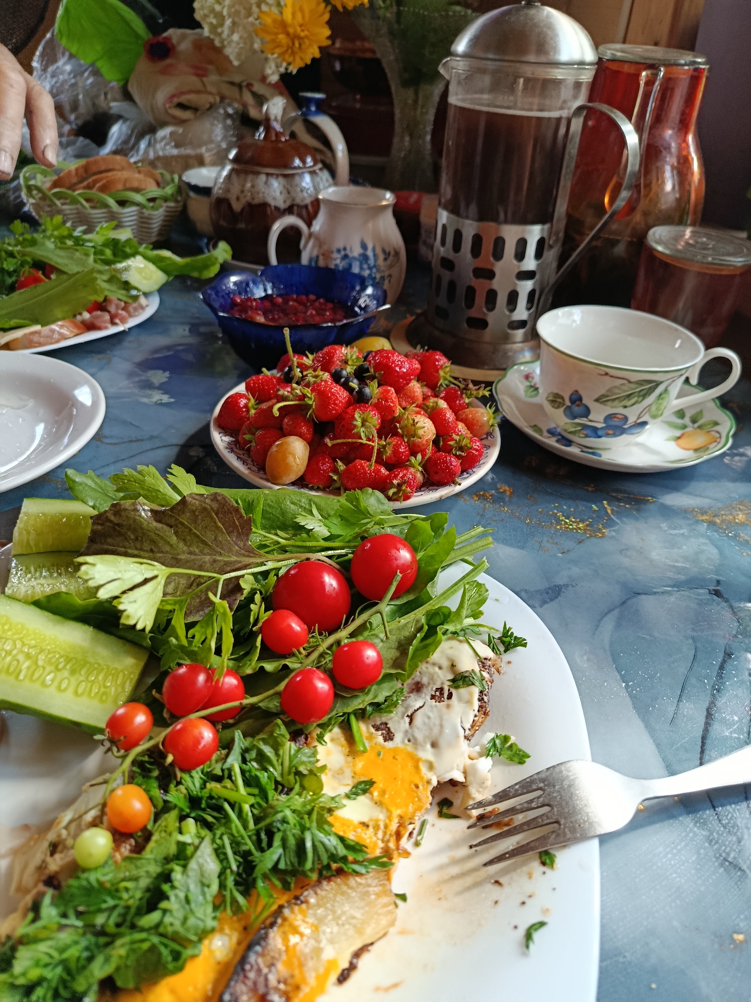 A close-up, high-angle shot captures a breakfast spread on a blue, textured surface, possibly a table. In the foreground, a white plate holds a meal consisting of a fried egg with a bright orange yolk, a piece of fish, and a garnish of fresh greens, cherry tomatoes, and cucumber slices. A fork rests on the plate, partially submerged in the food. To the right of the plate, a white teacup with a blue floral design sits on its saucer, near a glass French press filled with dark liquid, likely coffee. Several jars containing amber-colored liquid, possibly jam or preserves, and a small blue bowl filled with berries are also visible. In the background, a basket holds sliced bread, and decorative ceramic pieces add to the rustic aesthetic. A person's hand, with neatly trimmed nails, is visible in the upper left corner, reaching towards the bread. The overall setting suggests a casual, home-cooked breakfast.