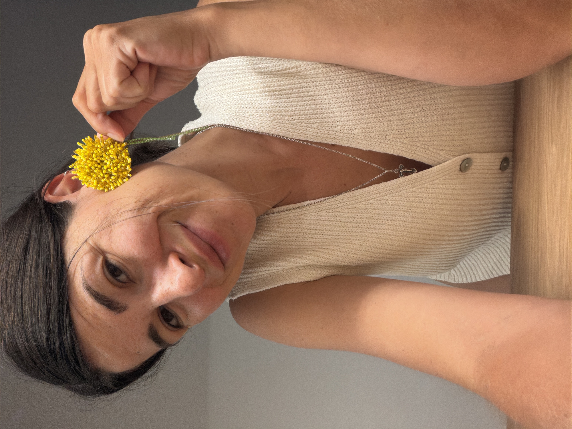A close-up, portrait-oriented photograph features a smiling woman in an indoor setting, possibly in Budva, Montenegro. She has dark hair and is looking directly at the viewer with a joyful expression. Her right hand is gently holding a vibrant yellow, spherical flower, composed of numerous small beads, positioning it near her left ear. A thin green stem is visible connecting to the flower.
The woman is wearing a cream-colored, V-neck knitted top with a ribbed texture and two light-colored buttons visible on the front. Around her neck, a delicate silver chain necklace with a small cross pendant is visible. The background consists of a plain grey wall on the left side and a warm, light-brown wooden surface on the right, suggesting a room interior. The lighting indicates a daytime setting. There is no visible text in the image.