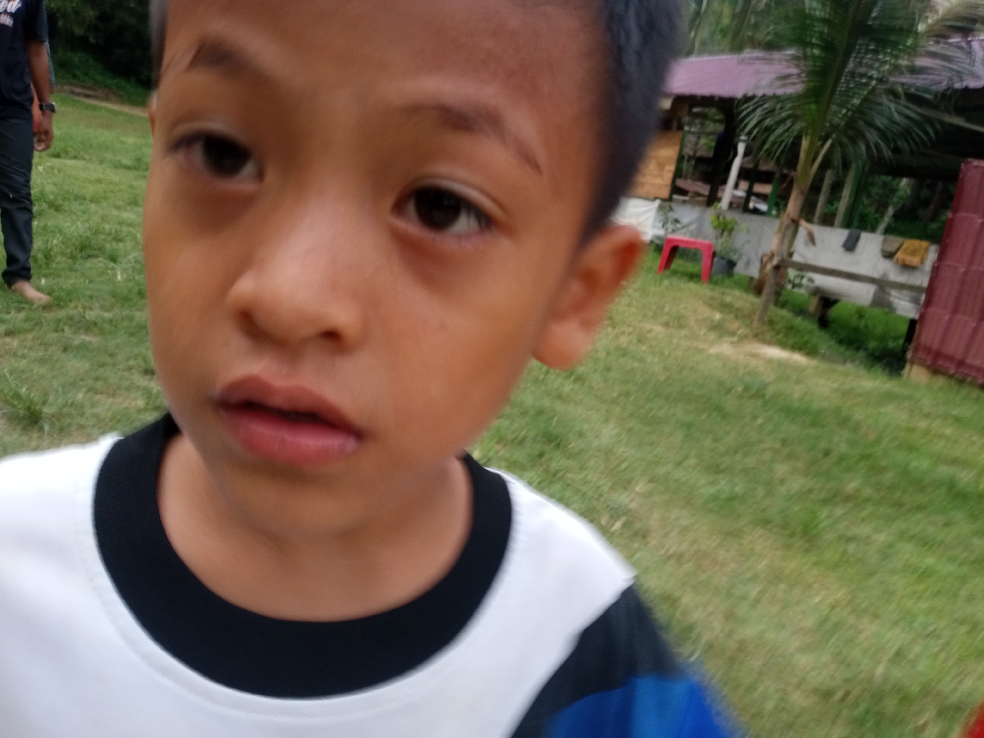 The image is a close-up, over-the-shoulder shot of a young boy with brown skin, dark hair, and brown eyes, wearing a white t-shirt with a black collar. His mouth is slightly open. In the background, a man wearing a dark shirt and dark pants, with his right foot bare, walks across a grassy area. Beyond the grassy area, there is a building with a red roof and palm trees. The scene appears to be outdoors in a tropical environment, consistent with Lhokseumawe, Indonesia.