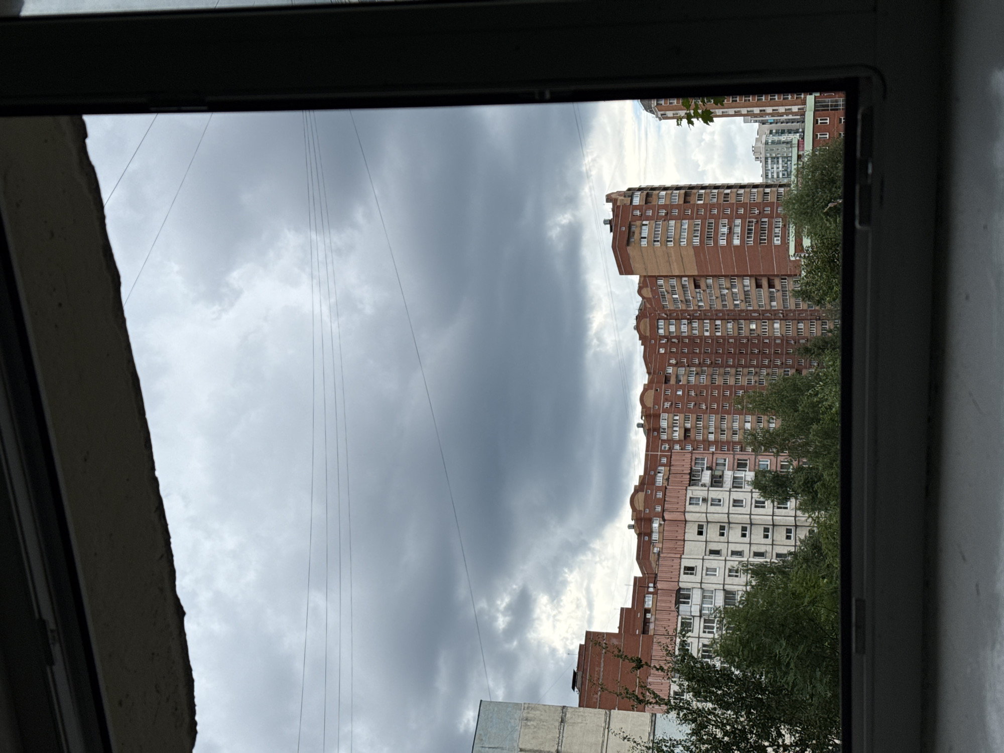 This is a photograph taken from a window, looking outwards at an urban landscape. The perspective is from within a darkened interior, suggesting the vantage point of someone inside a building, looking through a window frame. The frame itself is dark and partially visible, with a rough, unfinished edge on the left side of the image.
The scene outside is dominated by a dramatic sky filled with heavy, dark grey clouds, indicating overcast or stormy weather. Several thin, dark wires are strung across the sky, suggesting the presence of power lines or communication cables in the area.
In the background, a row of tall, multi-story buildings is visible. These buildings are primarily constructed with red brick and feature numerous windows, many of which appear to be light-colored. The architecture suggests a residential apartment complex. Trees with dense green foliage are interspersed between and in front of the buildings, adding a touch of nature to the urban setting.
The time of day appears to be daytime, but the overcast sky casts a dim, diffused light over the scene. The overall mood is somewhat somber due to the dark clouds. The location is identified as Khimki, Russia. There is no visible text in the image.