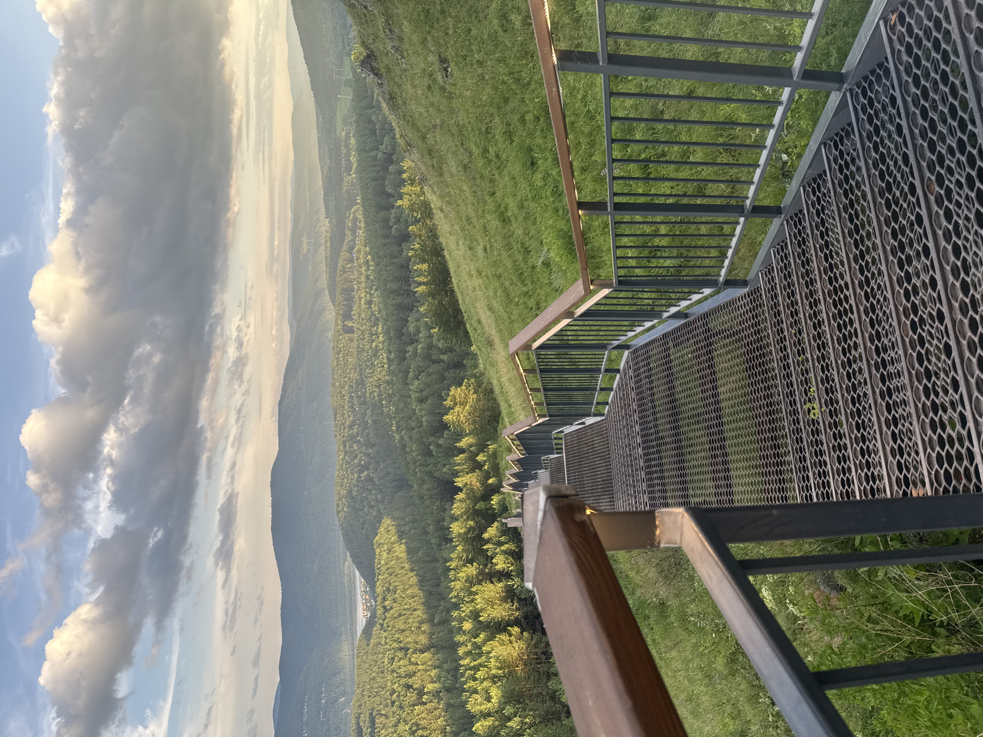 This is a vertical, eye-level shot taken from a metal staircase that curves down a grassy hillside. The staircase has metal railings and a grated metal surface. The scene overlooks a vast valley covered in lush green trees and rolling hills. In the distance, the landscape fades into a hazy blue due to atmospheric perspective. The sky on the left is dramatic, with large, illuminated clouds suggesting it is either sunrise or sunset. The lighting casts a warm glow on the clouds and the landscape. There are no people visible in the image. The location appears to be a scenic overlook or viewpoint, possibly in a mountainous or hilly region. The weather appears partly cloudy, with shafts of sunlight breaking through. The overall mood is serene and majestic, showcasing the natural beauty of the landscape.