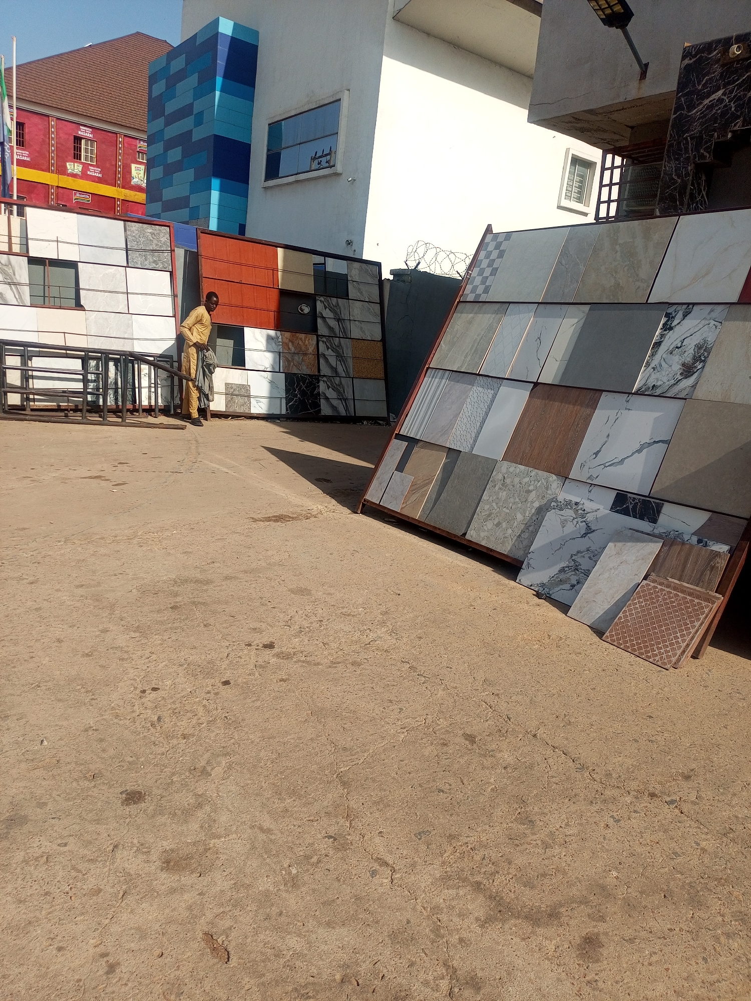 The photo shows a man standing near a display of tiles outdoors, potentially in a retail or market setting. The tiles are diverse in color and design, leaning against each other on the ground. The scene appears to be in a city, as suggested by the buildings in the background. A red building and another white building with blue colored bricks can be seen. The street appears to be a concrete surface. Maiburiji, Nigeria cannot be verified as the location.