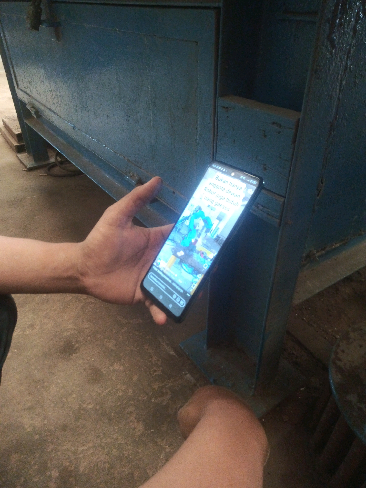 A person's hands are visible holding a smartphone, which displays text and an image. The text on the phone reads, "Bukan hanya anggota dewan. Robot juga butuh uang gaesss". The image on the phone appears to be a screenshot from a game or social media platform, featuring animated characters. The background is dominated by a large, blue, industrial-looking structure with a metal beam across the bottom. The ground is a rough, concrete-like surface. The lighting suggests an indoor or shaded outdoor environment. The overall scene does not provide explicit indicators of a specific location in Lebaniwaras, Indonesia.