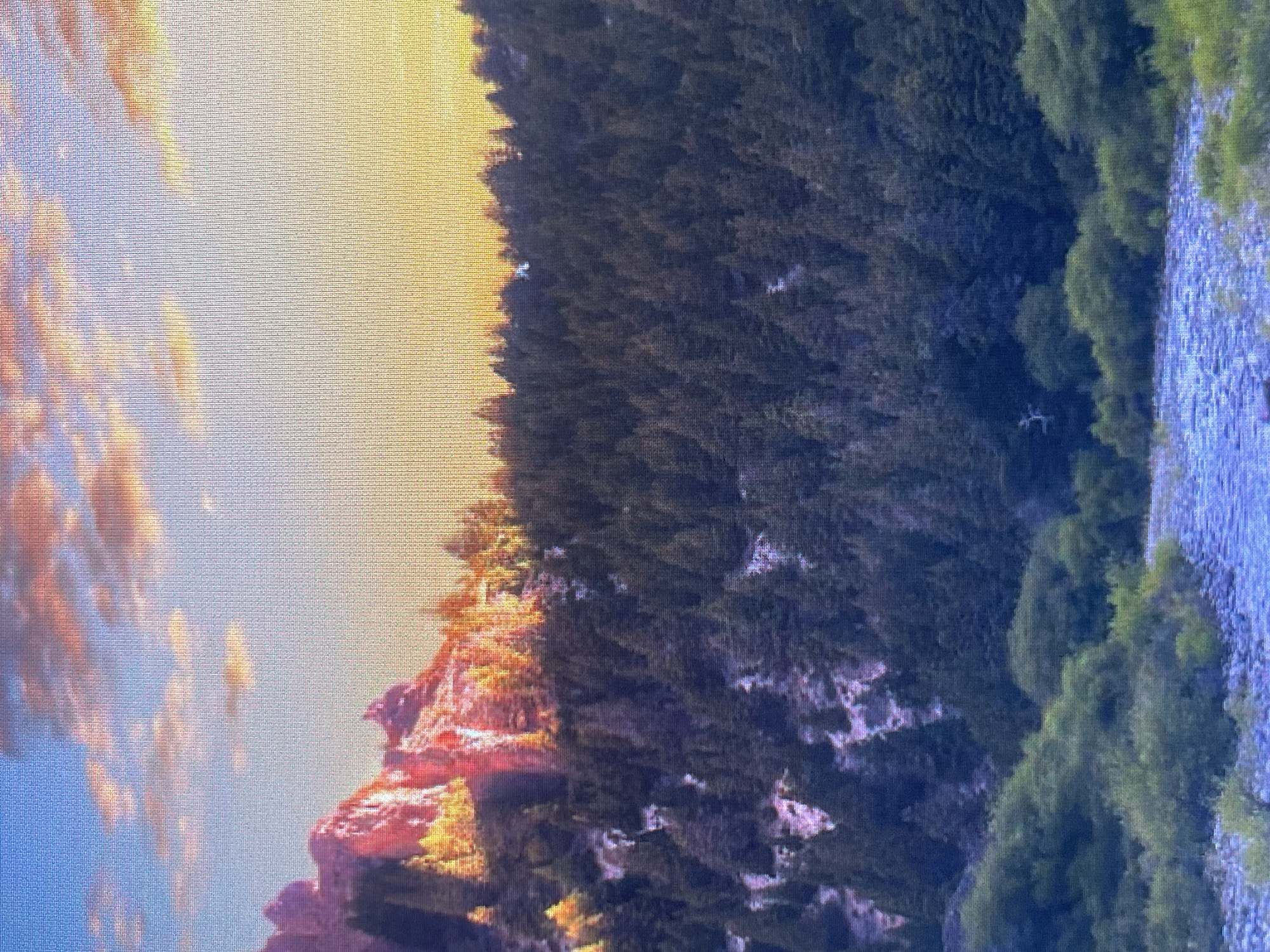 This is a landscape photograph taken at dusk. The image features a prominent mountain range on the left, illuminated by the warm, golden light of the setting sun. The mountains are covered in dense evergreen trees, with some patches of snow or light-colored rock visible. In the foreground, a winding river with clear, blue water flows through a rocky bed. Lush green vegetation lines the banks of the river. The sky above is a gradient of soft blues and oranges, with wispy clouds tinged with pink and yellow hues, indicative of sunset. The overall scene evokes a sense of tranquility and natural beauty. There are no people or man-made structures visible in the image, suggesting a remote, natural setting. The photograph is taken from an elevated viewpoint, looking down towards the river and across the forested mountains. The mention of Kafr Yasif, Israel is not supported by any visual evidence in the image itself.