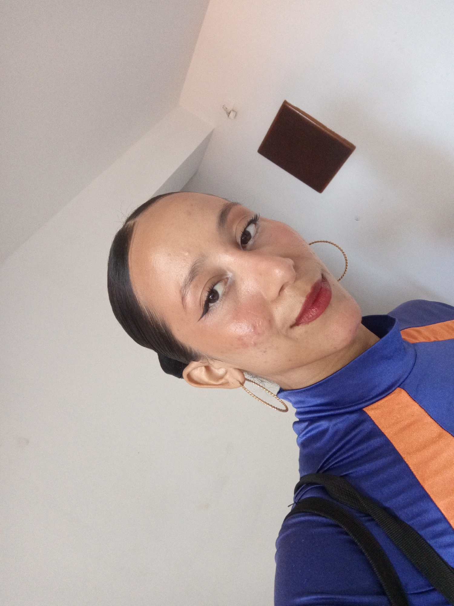 The image is a selfie taken indoors, seemingly in a room with white walls. A woman is the main subject. She has dark hair pulled back, dark eyeliner, and wears red lipstick. She is wearing a blue, high-necked garment with orange stripes. She is wearing gold hoop earrings. A brown, rectangular object is mounted high on the wall, likely a recessed light fixture. The location is unknown, the description of Porlamar, Venezuela cannot be determined from the image provided.
