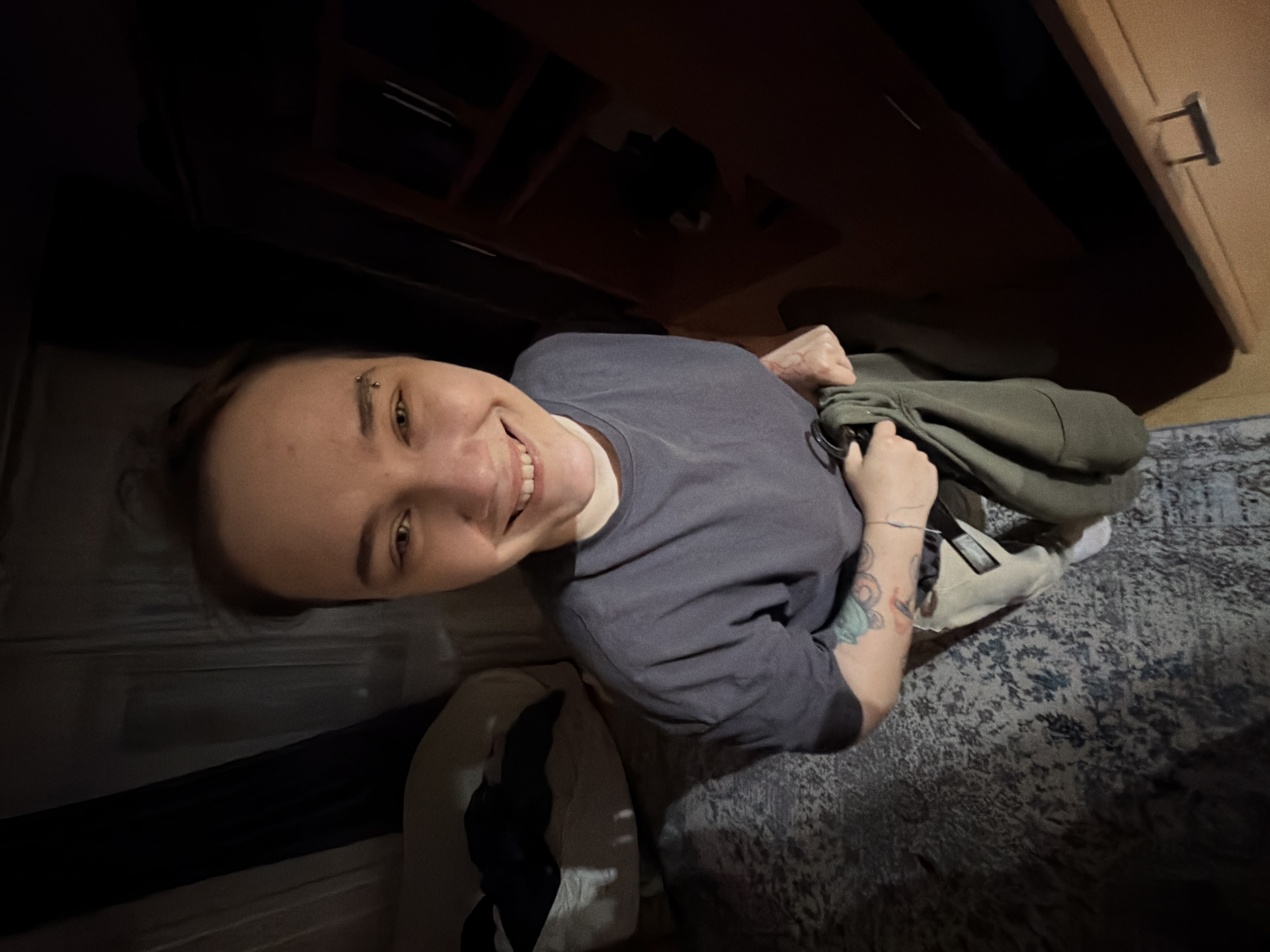 A fair-skinned person with short brown hair and a pierced eyebrow is smiling at the camera. The person is wearing a gray t-shirt and has tattoos on their left arm. They are holding a folded green hoodie. The person appears to be inside a room. A bed with dark covers is to the left. A dark cabinet above and to the right of the person is partially visible. There is a carpet with a dark floral pattern on the floor to the right of the person. A white bag with a black and white design sits behind the person. Based on the details available in the image, there is no specific evidence to suggest that it is located in the city of Chaikovsky, Russia. The precise location cannot be determined with the image provided.
