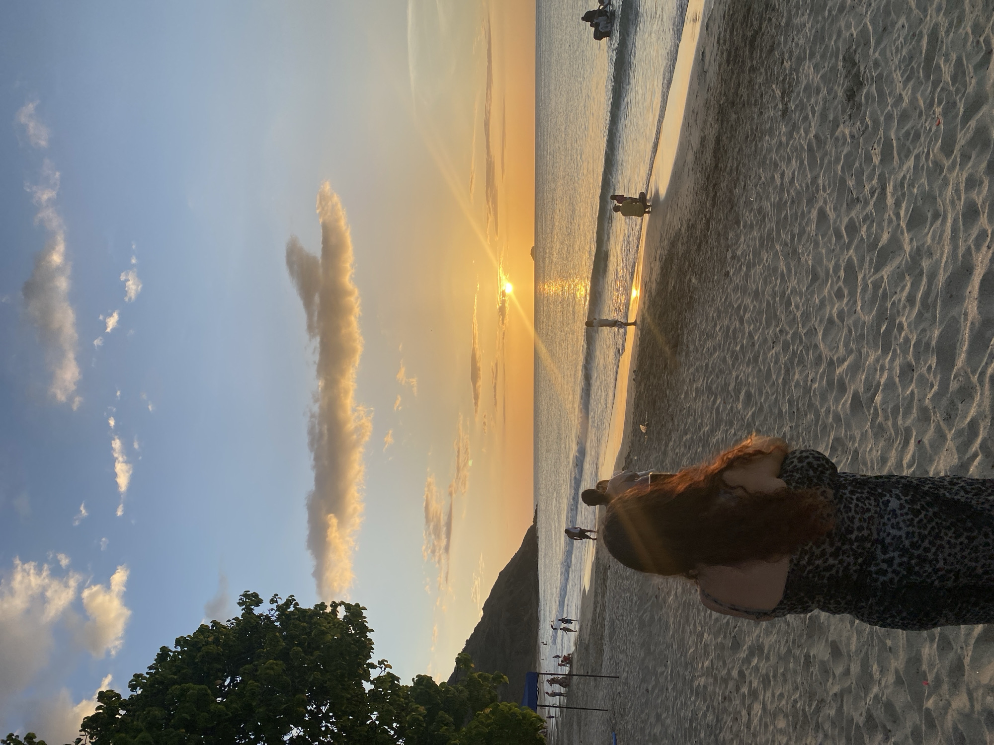 This is a horizontal, eye-level, medium shot of a beach at sunset. The sun is setting on the horizon, casting golden light across the ocean and sand. The sky is a mix of blue and orange, with scattered clouds. In the foreground, a woman with long, dark red hair is standing with her back to the camera, wearing a leopard print dress. In the middle ground, several people are walking on the beach and in the shallow water. The background shows a calm ocean with gentle waves and a small island in the distance. To the left, a lush green tree partially obscures the view of a dark, hilly landscape. The sand is light brown and textured with footprints and small indentations. This scene is taking place at Playa Redoma in Pedro González, Venezuela, known for its picturesque sunsets and tranquil atmosphere.