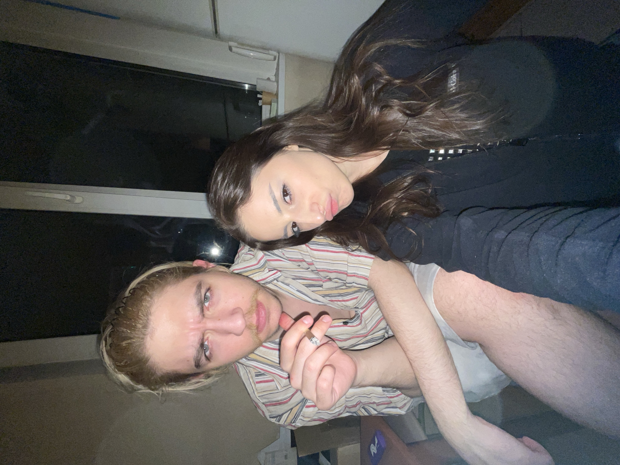 This media file presents a casual, close-up photograph of two young adults, a woman and a man, taken indoors during what appears to be the evening or night in Moscow, Russia. The image is rotated approximately 90 degrees counter-clockwise.
The woman is positioned behind and slightly above the man, her long, dark, wavy hair cascading around her. She wears a black long-sleeved top or hoodie, on which a "Calvin Klein" logo is partially visible on the left chest. She gazes directly at the camera with a subtle pouting expression and dark, made-up eyes.
In the foreground, the man wears a short-sleeved shirt featuring horizontal stripes in red, yellow, white, and black. He has light blonde or dirty blonde hair, styled back with a dark hairband, and light-colored eyes. He sports a light mustache and a small patch of hair on his chin. He is holding a lit cigarette between the thumb and index finger of his right hand, adorned with a ring on the ring finger, and maintains a direct, intense stare at the camera.
The background is dimly lit, revealing a dark window on the upper left, which reflects some ambient light, and a lighter-colored wall or ceiling above it. Indistinct details, possibly furniture or boxes, and a section of the man's hairy leg are visible in the lower right portion of the frame. The overall impression is that of a candid, intimate snapshot in a relaxed, low-light indoor setting.