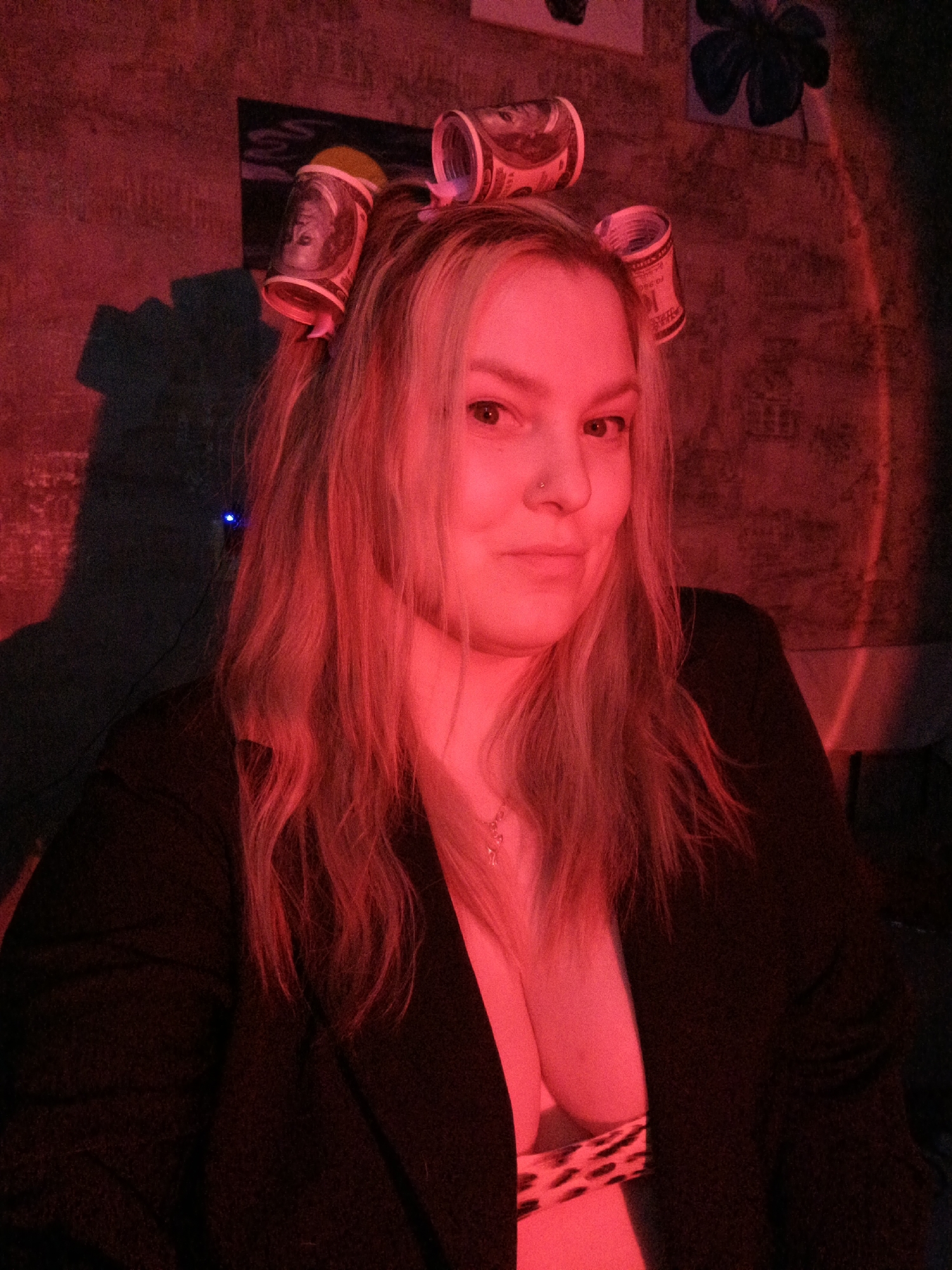 A woman with blonde hair and a nose ring is illuminated by a red light. She is wearing a black blazer, revealing a leopard-print top underneath. Her hair is styled with curlers made from what appears to be rolled-up US dollar bills. The background includes dimly lit walls, with a dark floral painting visible on the upper right. A small blue light is also visible on the wall behind her. The location is not discernible as Марковский, Russia from this image.