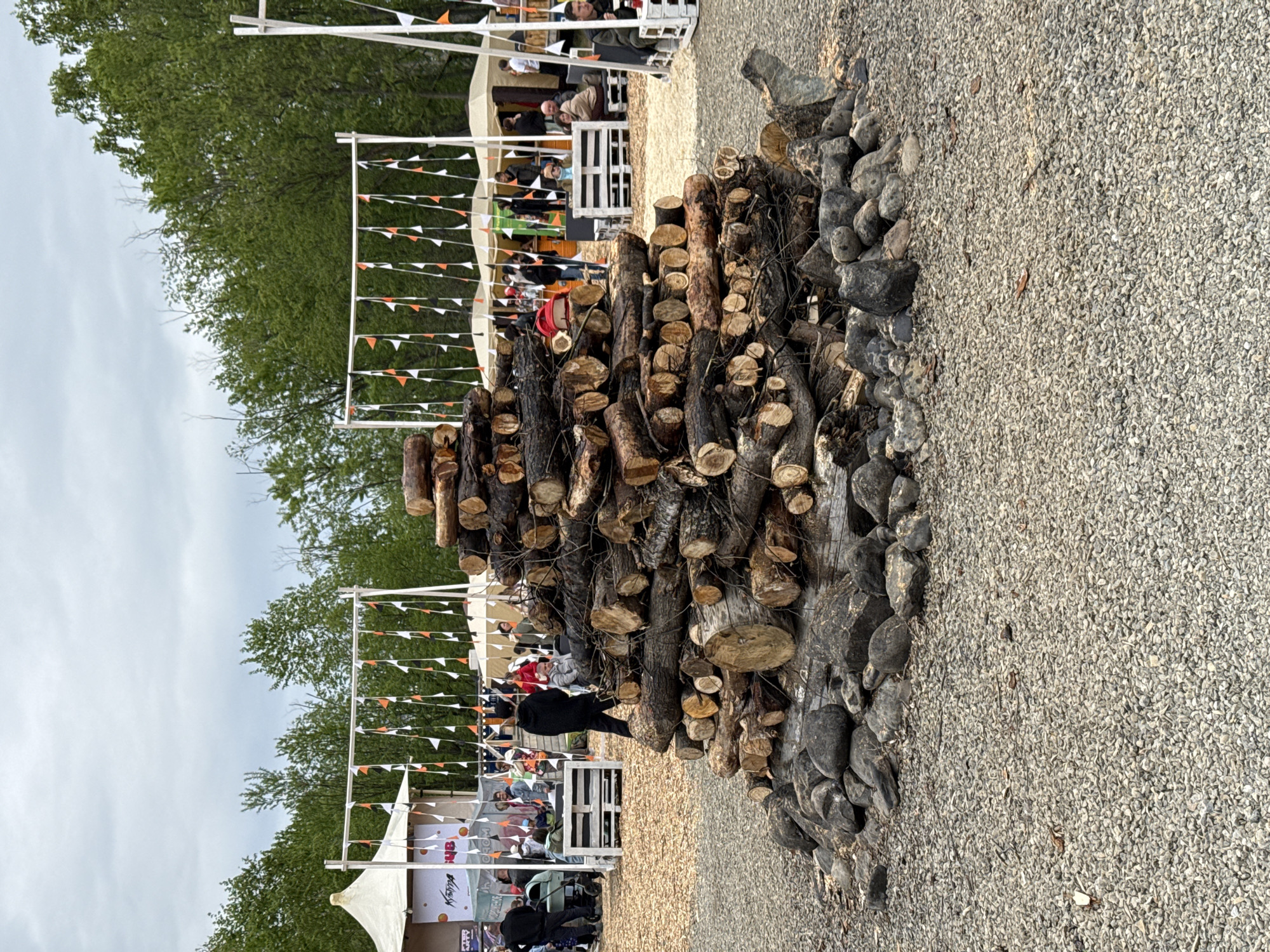 The image depicts an outdoor gathering or festival taking place in Paratunka, Russia, with a focus on a large, centrally located bonfire pile. The bonfire is constructed from a substantial quantity of chopped logs, artfully stacked and surrounded by dark-colored stones at its base. In the background, white tents are set up, suggesting stalls or service areas for the event. Rows of small, colorful triangular flags, predominantly orange and white, are strung between white poles, decorating the area. Several people are visible in the background, engaged in various activities such as walking, sitting, and standing near the tents. The environment appears to be an open space with trees lining the perimeter, indicating a semi-rural or park-like setting. The ground is covered with gravel and wood chips. The sky is overcast with clouds.
