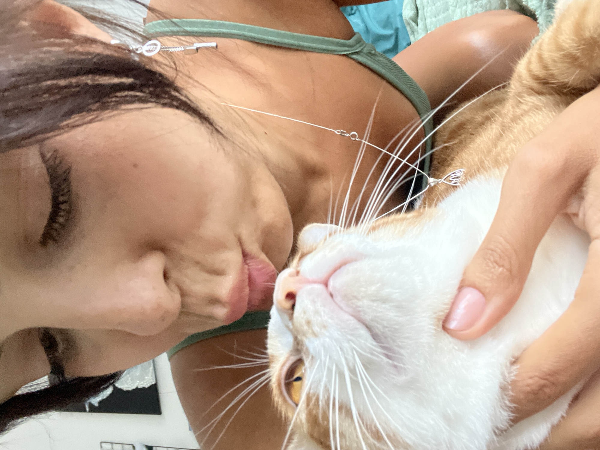 A woman with dark hair and tan skin kisses a white and orange tabby cat. The woman wears a green tank top and a silver necklace. The location is unknown, including whether it is in Kuta Selatan, Indonesia.
