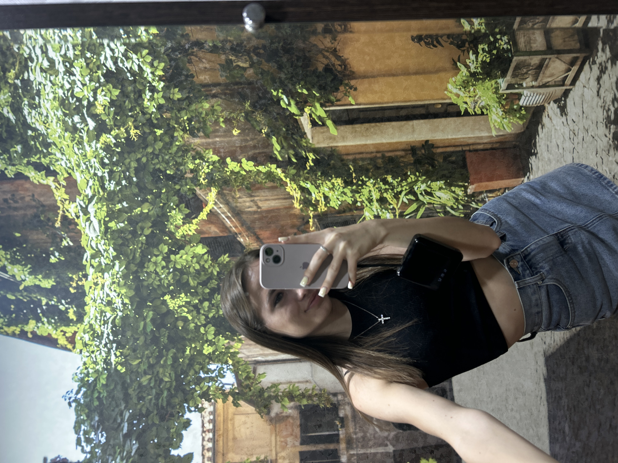 A young adult female is depicted taking a mirror selfie indoors. She has long, straight, dark brown hair and is wearing a black sleeveless crop top, a blue denim mini skirt, a silver-colored cross pendant necklace, and a silver ring on her left hand. She is holding a light purple/lavender smartphone with a triple camera system in her left hand, and a small, black rectangular object in her right hand below the phone. Her visible fingernails are painted a pale yellow-green. The background is a large mural or print depicting an old European street scene. This scene features buildings with ochre and brown walls, abundant green ivy and climbing plants covering the walls, particularly in the upper and right sections. Potted plants and wooden planters with greenery are visible at ground level. The ground is paved with cobblestones, and a narrow pathway is visible. A silver circular knob is present on the right edge of the frame, suggesting a mirror frame or door. The actual location is an unknown indoor setting, in the city of Unknown, Unknown.