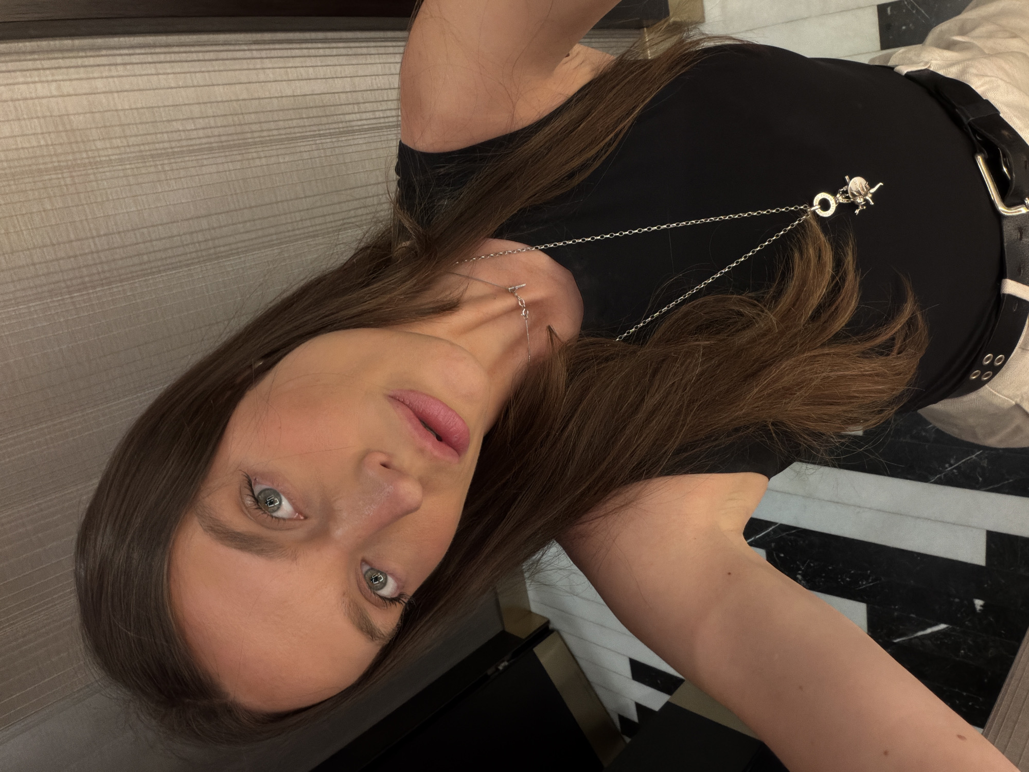 A young woman with long, brown hair and blue eyes is taking a selfie. She is wearing a black tank top and a silver necklace with a pendant. Her skin has a rosy hue, and her lips are painted pink. The background appears to be an indoor setting with a patterned floor and a textured wall. The woman is looking towards the camera with a slight tilt of her head. The composition of the image is a close-up, focusing on her face and upper body.