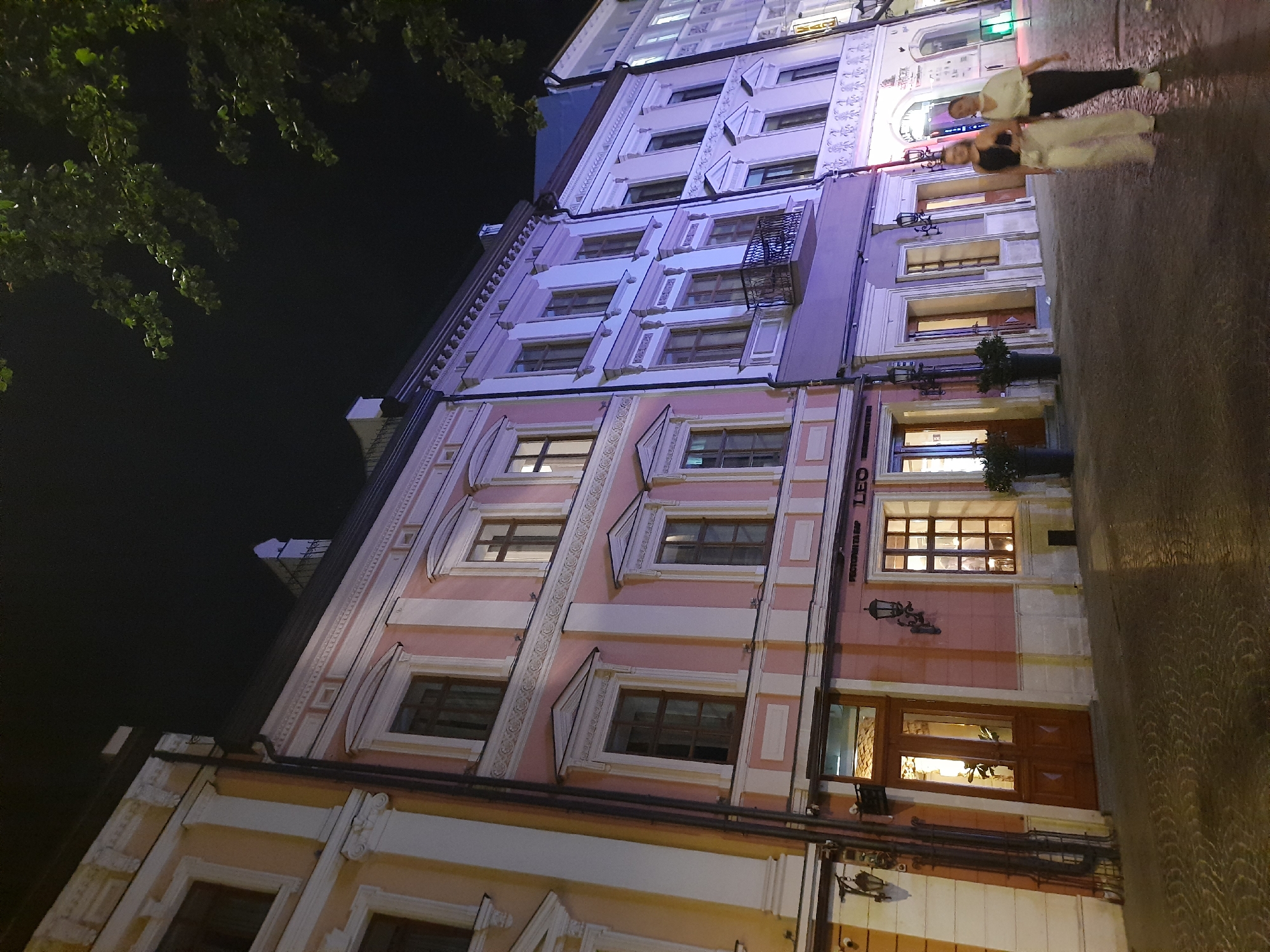 The image captures a nighttime scene on a European-style street in Lviv, Ukraine. Two young women, one in a black top and white pants, and the other in a white t-shirt and dark pants, are walking away from the viewer. They are on a cobblestone street reflecting the lights from the surrounding buildings. The buildings themselves are multi-story structures with ornate architectural details, featuring large windows and decorative cornices. The facades are painted in pastel colors, predominantly pink and light purple, with accents of white and yellow. Some windows are illuminated, suggesting interiors are active. A tree with green foliage is visible in the upper right corner, its leaves silhouetted against the dark night sky. The overall impression is of a lively urban environment at night, illuminated by artificial light.