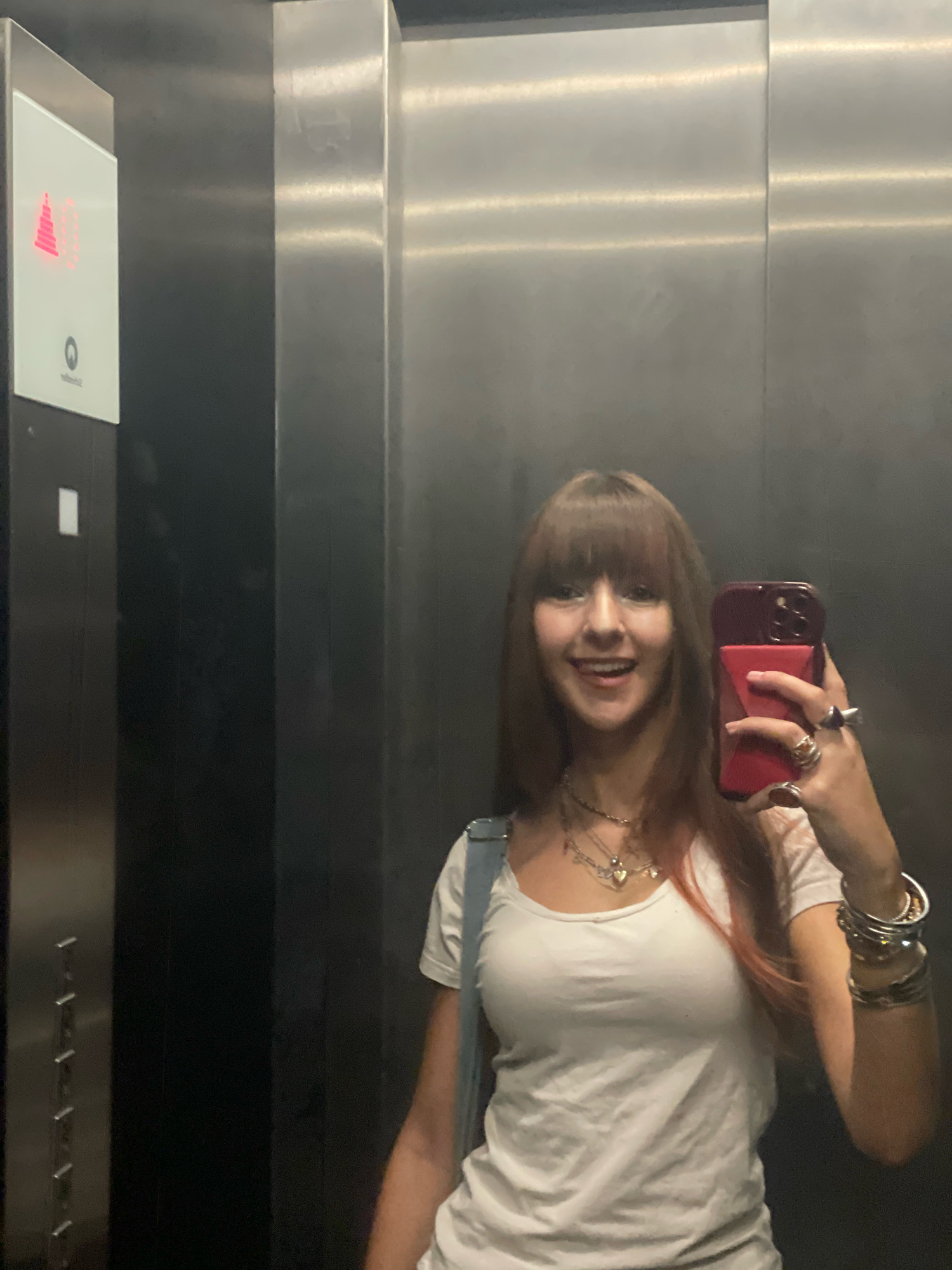 A young woman with long, reddish-brown hair and bangs takes a selfie in an elevator. She is wearing a white t-shirt, a denim strap, and several necklaces and bracelets. She is holding a red-cased smartphone in her right hand, which is adorned with rings. Her left hand is also visible, with fingers extended and nails painted. The elevator interior is metallic, with vertical seams visible on the walls. To the left of the woman, an illuminated panel displays a red graphic and a circular button. The scene is consistent with an elevator in a modern building, potentially located in Raanana, Israel, given the context provided.