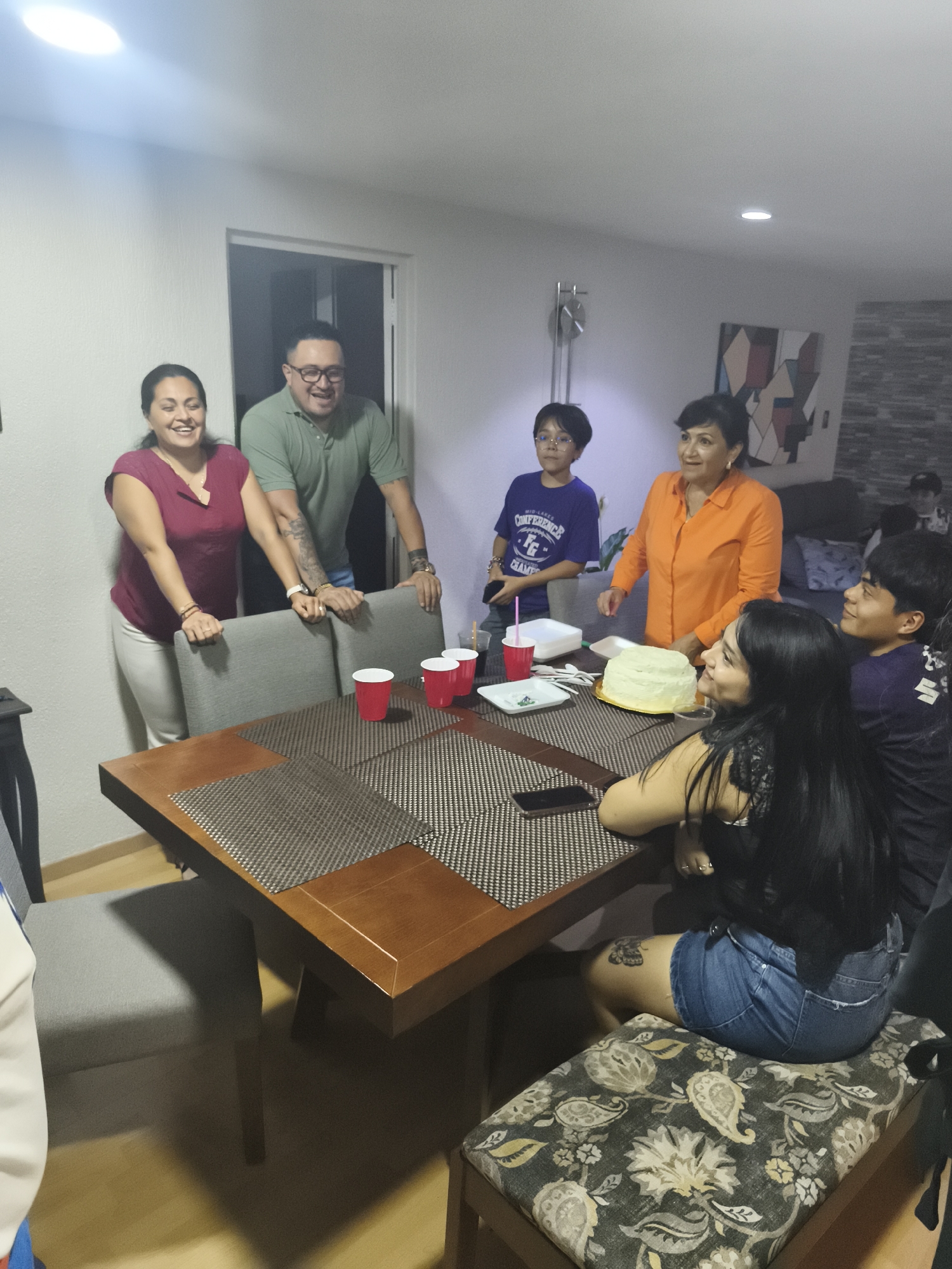 A group of people is gathered around a table in what appears to be a home setting. A woman in a maroon top and white pants stands to the left of the table, next to a man wearing glasses and a green shirt. Both have their hands on the backs of chairs and appear to be smiling. A younger boy with glasses, wearing a purple shirt, stands behind the table to the left, and an older woman in an orange blouse is next to him, with her arm extended towards the table. A partially visible cake sits on the table, along with red cups, plates, and cutlery. Two people are seated at the table: a woman with long black hair and denim shorts, and a boy in a dark shirt. A cell phone rests on the table.
The room is lit by overhead lighting, and there is a painting or art piece on the wall.
There is insufficient information to identify the location as Mexico City, Mexico.