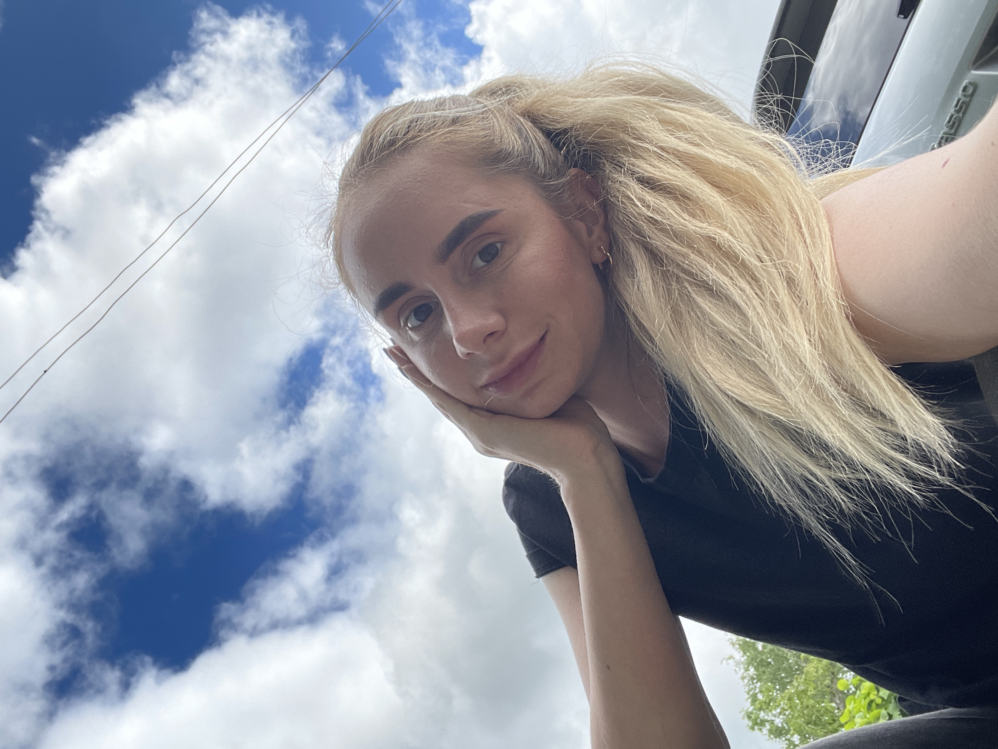 The image displays a low-angle selfie of a woman outdoors in Yasnoe, Russia. The primary subject is a woman with blonde hair, fair skin, and dark eyes, looking directly at the camera with a subtle smile. Her head is tilted slightly to the viewer's left, and her right hand is positioned on the side of her face. She is wearing a black t-shirt, and her blonde hair is pulled back in a ponytail with some loose strands framing her face. Small hoop earrings are visible in both ears.
The background consists of a bright blue sky with numerous scattered white cumulus clouds. Two thin, dark lines, resembling power lines or cables, traverse horizontally across the upper right portion of the sky. In the lower-left background, green foliage is visible, indicating trees or bushes. To the lower-right, the rear section of a dark-colored vehicle is partially visible, including a dark window and the chrome lettering "PASSO" on the vehicle's body, identifying it as a Toyota Passo. The scene is illuminated by natural daylight.