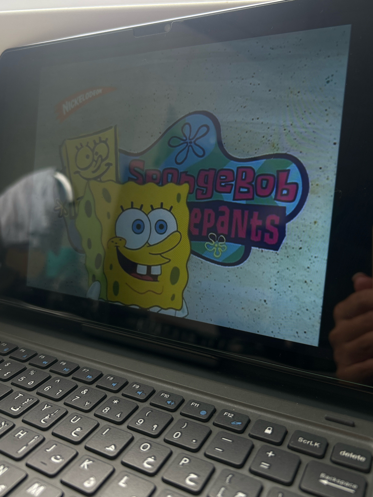 A Spongebob Squarepants logo is displayed on a laptop screen. The logo features Spongebob himself on the left, with the words "Spongebob" in blue and pink lettering above "pants" in pink lettering. A Nickelodeon logo is visible in the top left corner of the screen. The laptop keyboard, which includes Arabic lettering on some keys, is in the foreground. The setting is indoors, with the laptop on a surface, and a hand is visible in the reflection on the screen. The image is taken from a slightly elevated angle, looking down at the laptop. There is no visible information within the image that indicates the location is Khyrdalan, Azerbaijan.