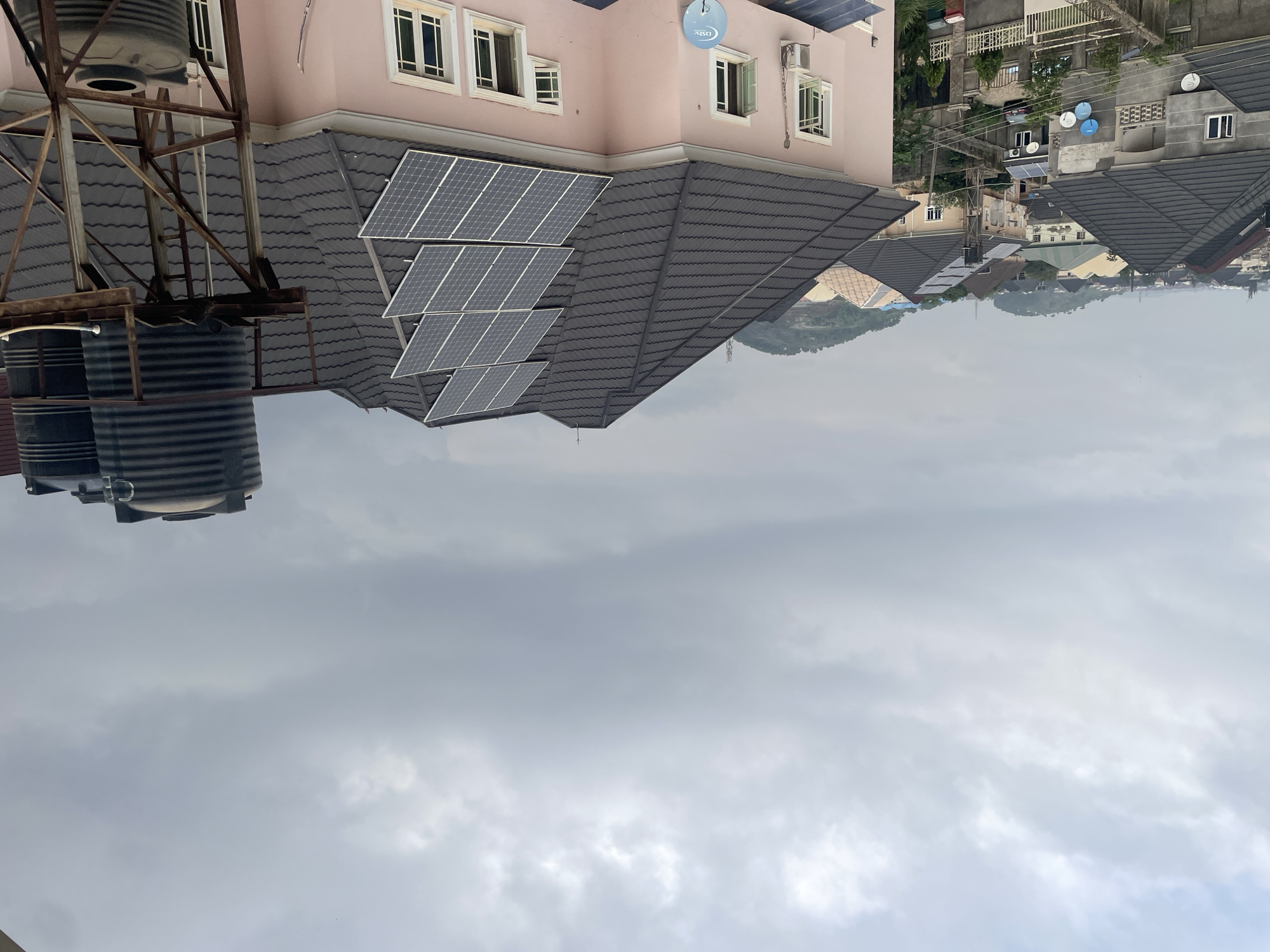 This is a high-angle exterior shot of a residential area in Abuja, Nigeria, under a cloudy sky. The foreground is dominated by the dark, tiled roof of a pink house, on which a bank of six solar panels is installed. To the right of this house, elevated on a rusted metal platform, are two large black water tanks, with a third smaller tank visible below.
Beyond this pink house, other houses with similarly dark tiled roofs are visible, some with their own solar panels and satellite dishes. The architecture suggests a modern residential development. In the background, rolling hills and more houses extend towards the horizon, partially obscured by the overcast weather. The overall impression is of a suburban or semi-urban environment in Abuja, with a focus on sustainable energy solutions like solar panels and rainwater harvesting. No people are visible in the image.