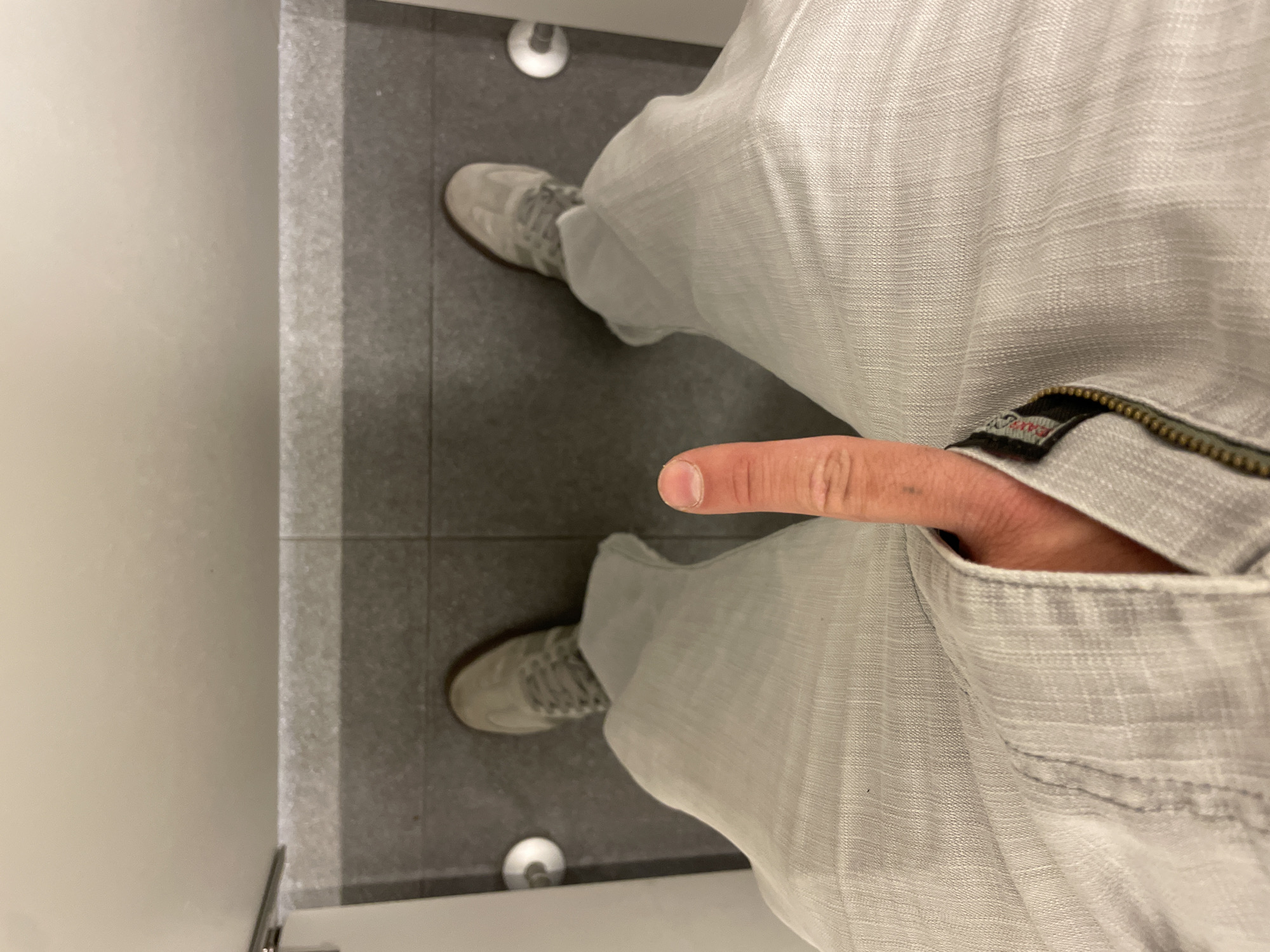 The photograph depicts a person standing in a bathroom with gray tile flooring. The individual is wearing gray pants and light gray shoes. A finger is protruding from a pocket on the right side of the pants. The environment consists of a bathroom stall with a white wall partially visible on the left. The location is Unknown, Unknown.
