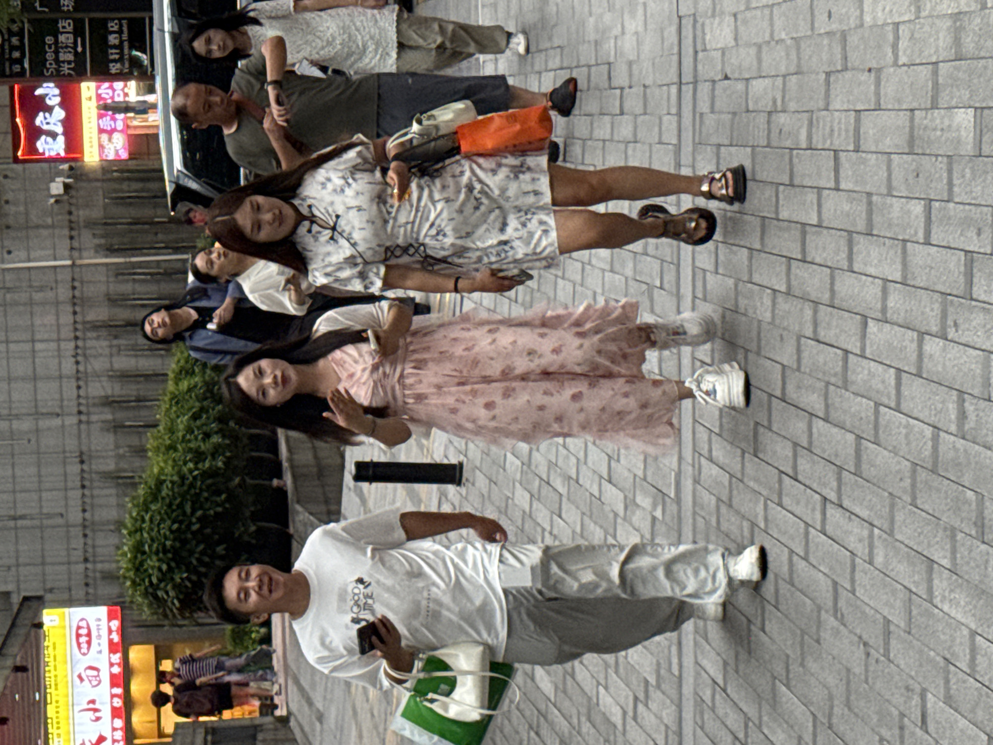 In Yuzhong District, China, on a paved sidewalk, three individuals are walking. In the foreground, a man in a white t-shirt and light-colored pants holds a smartphone and a shopping bag. To his right, a woman in a pink floral dress walks alongside him. Behind them, a woman in a white and blue floral dress carries shopping bags. The background reveals buildings with neon signs, a bus, and lush green bushes. The scene captures a moment of urban pedestrian activity.