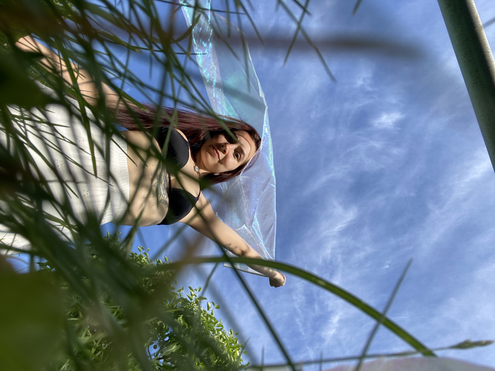 The image presents a unique low-angle view looking upwards through blurred green foliage towards a young woman against a bright blue sky. The woman, with long reddish-brown hair, is positioned diagonally in the frame. She wears a black bra or bikini top and a white, possibly embroidered, skirt or sarong, with tattoos visible on her left arm. She holds a translucent, iridescent fabric or scarf, which catches the light and displays subtle shifts in color, extending it across the upper right portion of the image. Her gaze is directed towards the camera, and she has a gentle, serene expression.
The background is dominated by a clear, vivid blue sky with sparse, wispy white clouds, indicating a sunny day, likely late morning or afternoon. The foreground features various green leaves and blades of grass, heavily blurred, framing the central subject and creating a sense of depth. On the far right, a narrow, light-colored, cylindrical object, possibly a pole or pillar, is partially visible. The scene suggests an outdoor setting, such as a park or garden, in Chaikovsky, Russia, with the woman enjoying the bright, clear weather. No text is visible in the image.