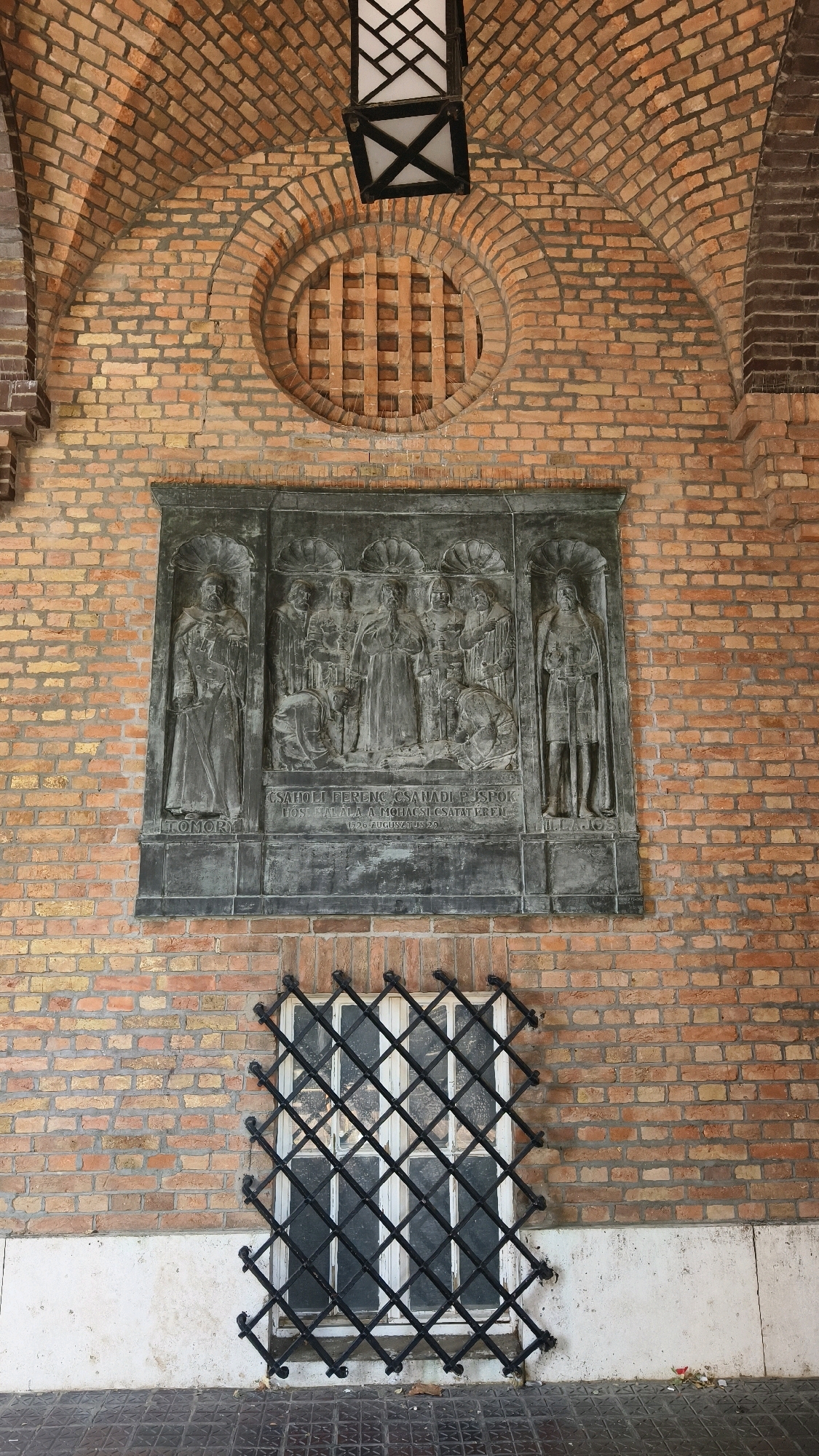 The image shows a bronze relief plaque mounted on a brick wall. The plaque depicts a central scene with several figures, including one kneeling figure. Flanking this central scene are two standing figures in architectural niches. Below the figures, an inscription is visible. Above the plaque, there is a circular brick pattern with a grid-like structure. A modern wrought-iron security grille covers a window below the plaque, set against the brick wall. The architecture suggests an outdoor or semi-outdoor setting, possibly a courtyard or portico. The plaque is located at the entrance of the Dóm (Cathedral) in Szeged, Hungary. The inscription on the plaque reads "CSAHOLI FERENC CSANADI PUSPÖK HOSE HALALA A MOHACSI CSATAT EREN. 1526 AUGUSZTUS 29." This commemorates the death of Bishop Ferenc Csaholi in the Battle of Mohács on August 29, 1526.