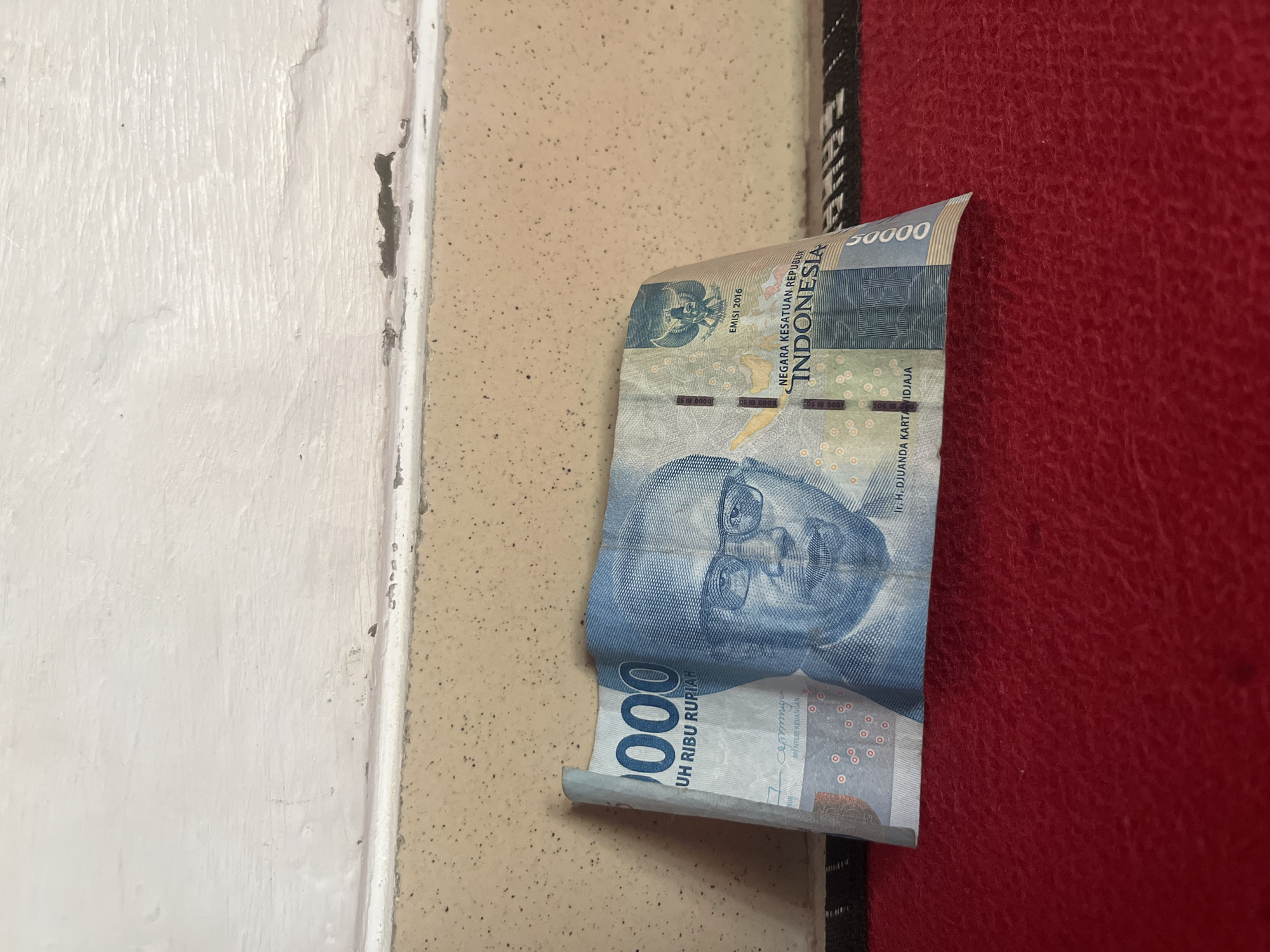 A folded Indonesian 50,000 rupiah banknote is placed on a red textured surface. The banknote is predominantly blue and features the portrait of Djuanda Kartawidjaja. The denomination "50000" is visible on the right side of the banknote. The background consists of a light-colored wall with a textured surface and some peeling paint near the top edge. The context provided states the location is Glagah Kulon, Indonesia.
