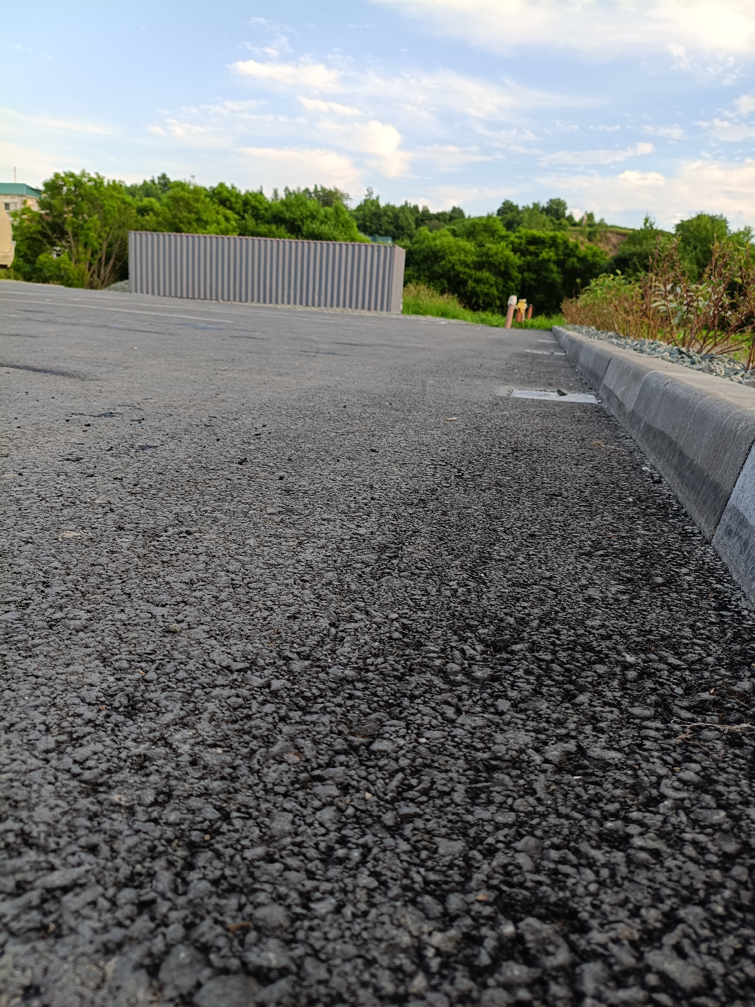 The image is a low-angle, ground-level shot of an asphalt surface with a concrete curb running along the right edge. The asphalt is dark gray and appears to be slightly wet or recently paved. The curb is gray and textured, with a slight sheen, indicating it may also be damp. Beyond the curb, a strip of gravel and then some dry, reddish-brown foliage is visible. Further back, a lush green line of trees and bushes forms a backdrop, interspersed with taller trees and a distant hillside. To the left, a grey corrugated metal fence is partially visible, and behind it, a building with a green roof can be seen. The sky is blue with scattered white clouds. Two orange traffic cones and a white utility box are positioned near the edge of the asphalt in the distance. The scene appears to be an outdoor urban or semi-urban environment, possibly a road or parking area, in Korsakov, Russia, given the presence of the specific building and the general landscape. No people are visible in the image.