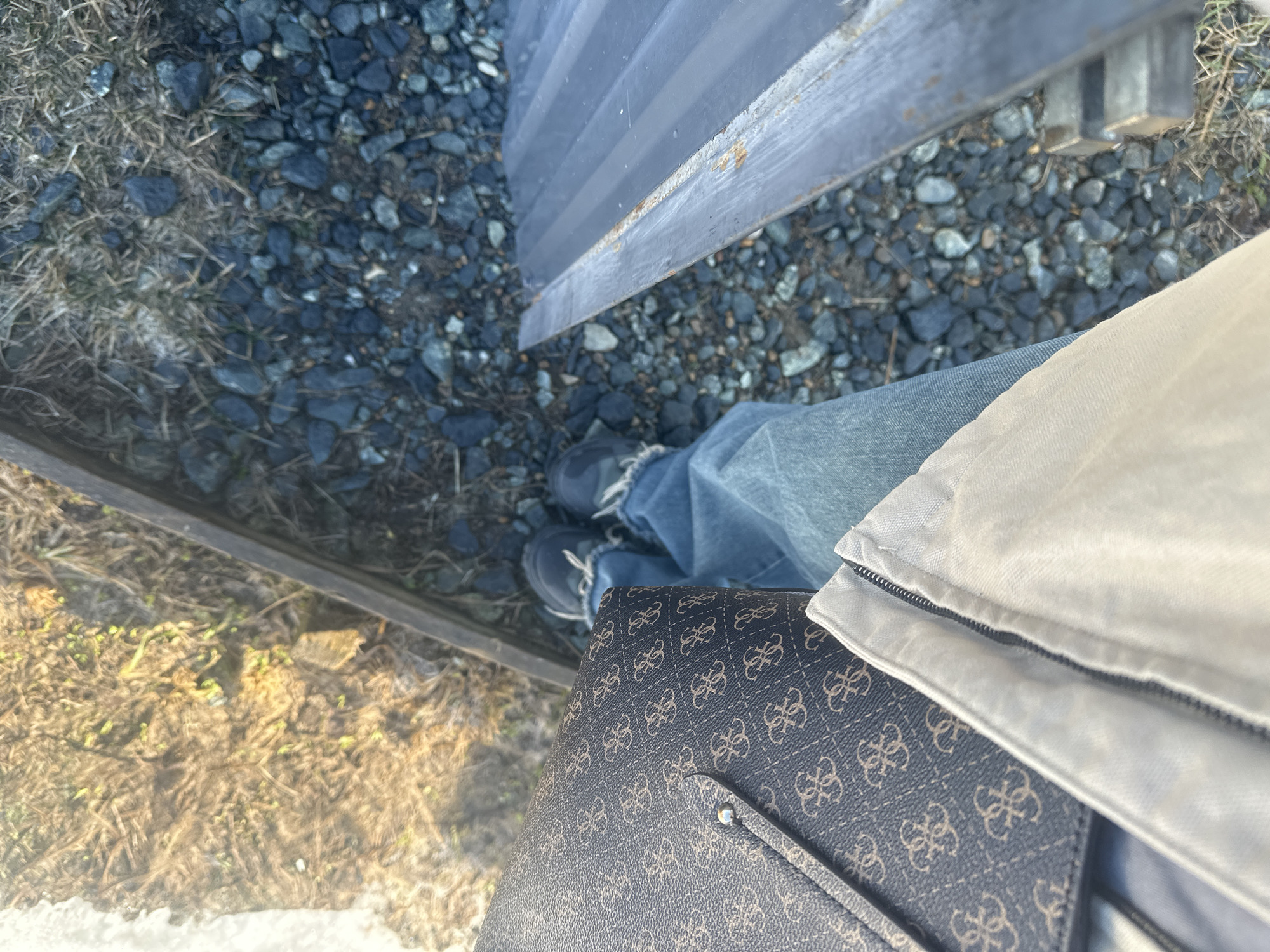 The image shows a first-person perspective looking down at the ground, possibly taken in an outdoor environment. The ground consists of gravel and patches of dry grass or vegetation. There are pieces of wooden planks or structures on the left and top of the frame, possibly forming a walkway or a low fence. A person's legs, clad in denim jeans and dark sneakers, are visible. A brown patterned purse or bag rests against the person's body. A beige or white jacket is visible along the bag's top edge. The location is described as Kharnavy, Russia; however, there is no specific element in the image that corroborates the exact location, it only shows general outdoor elements. There is no visible interaction or actions taking place.