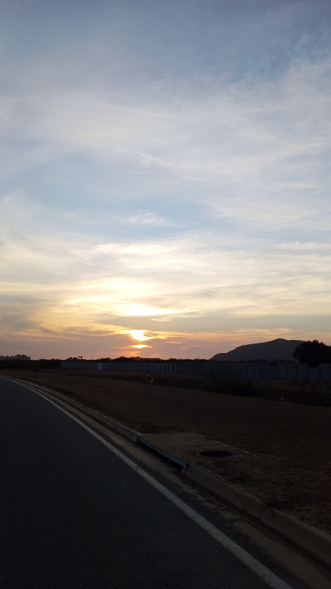 The image captures a serene sunset landscape, likely from the perspective of a vehicle or pedestrian on the side of a road. The sky dominates the upper two-thirds of the frame, showcasing a beautiful blend of colors. Near the horizon, the sun, partially obscured by clouds, casts a brilliant golden-orange glow across the sky, with hues of pink and soft yellow blending into wispy, elongated clouds. Higher up, the sky transitions to a lighter blue with delicate white cirrus clouds stretching across.
In the foreground, a dark asphalt road or highway curves gently to the left, marked by a solid white line along its edge. To the right of the road, a concrete curb and a drainage ditch run parallel. The ground immediately adjacent to the road is dark brown, appearing dry and somewhat barren, with patches of sparse vegetation.
In the midground, a long, low, light-colored barrier wall or fence stretches horizontally across the scene, separating the road area from the distant landscape. Beyond this wall, the ground remains dark and flat, possibly undeveloped land. On the far left horizon, faint silhouettes of buildings or structures are visible. On the right, a prominent dark mountain or hill range rises against the colorful sky, its silhouette appearing robust and irregular. Some dark trees are discernible near the base of the mountain.
The time of day is clearly sunset, as indicated by the vivid colors near the horizon and the deep shadows cast over the landscape elements. The weather appears calm with scattered clouds. There are no people, animals, or discernible text visible in the frame. The overall mood is peaceful and expansive.