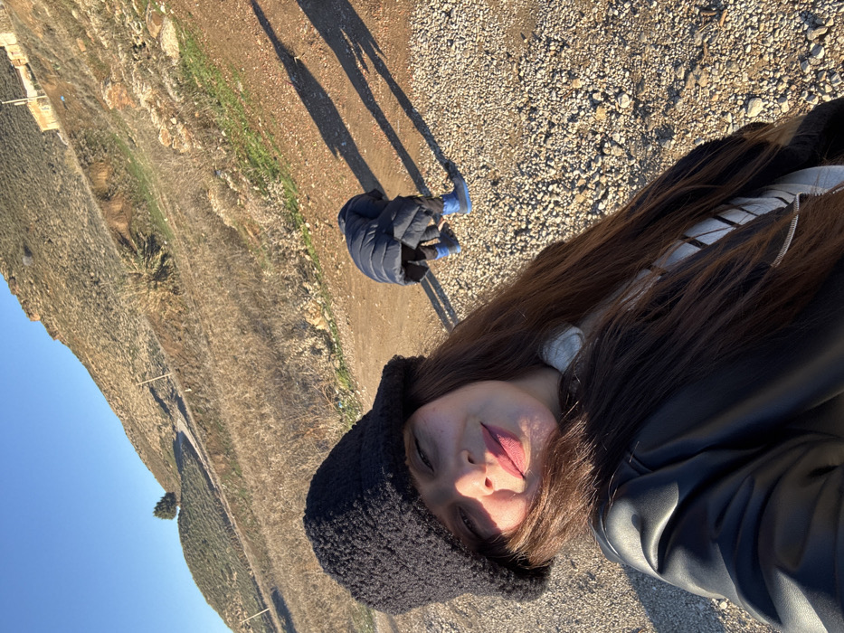 The image captures an outdoor scene in Azrou, Morocco, under clear, direct sunlight, evident from prominent shadows stretching across the ground. In the immediate foreground, a female with long brown hair, wearing a black textured bucket hat, a light-colored striped top, and a black leather-like jacket, smiles directly at the camera, indicating a selfie. Her face is brightly illuminated from the left. In the midground, to the right of the primary subject, a second individual, likely a child, is bent over on a gravel and dirt path, facing away from the camera. This individual wears a dark blue puffer jacket and bright blue boots. The background features a large, ascending hillside, predominantly covered in dry, brown vegetation with sparse green patches. A distinct evergreen tree is visible on a minor peak on the left side of the hillside, and faint structures are discernible higher up on the main slope. The sky above is clear and blue.